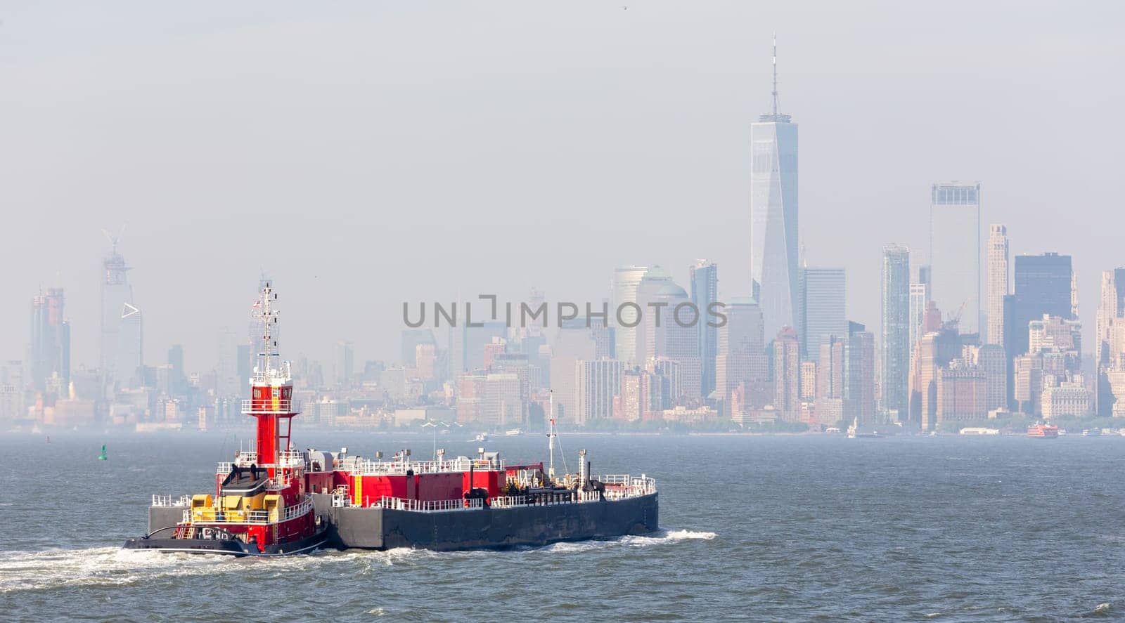 Freight tug pushing cargo ship to the port in New York City and Lower Manhattan skyscarpers skyline in background. New York City, USA.
