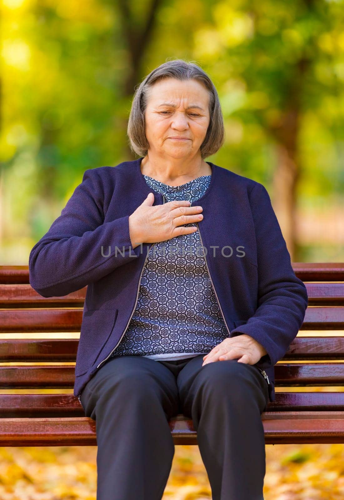 Senior lady clutching her chest in pain at the first signs of angina or a myocardial infarct or heart attack, upper body in park on a autumn day