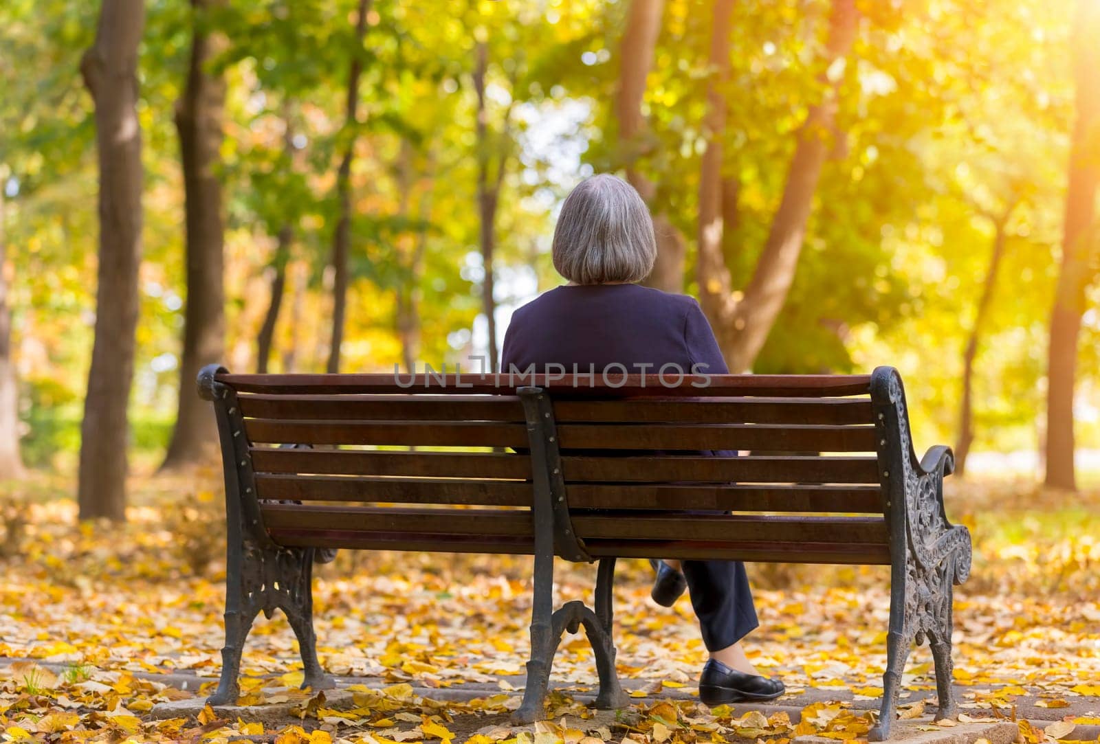 Elderly woman sitting on a bench in autumn park. Lonely aged woman sitting on bench in autumn park lookes at trees.