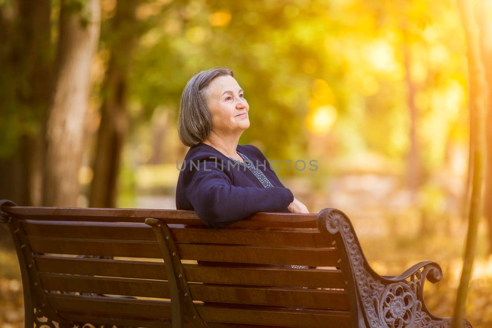 Elderly woman sitting on a bench in autumn park. Aged woman sitting on bench in autumn park looks and smiling at the sun.