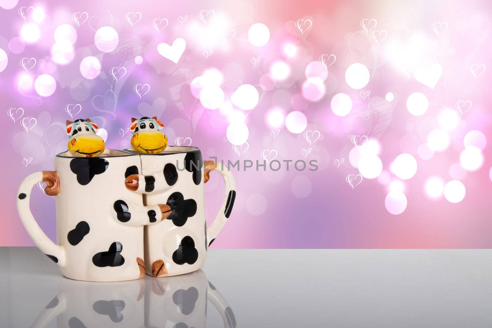 Valentine day concept. Sweet cows hug each other as a form of two mugs on a bright table in front of a love hearts bokeh background. Template for your display product montage.