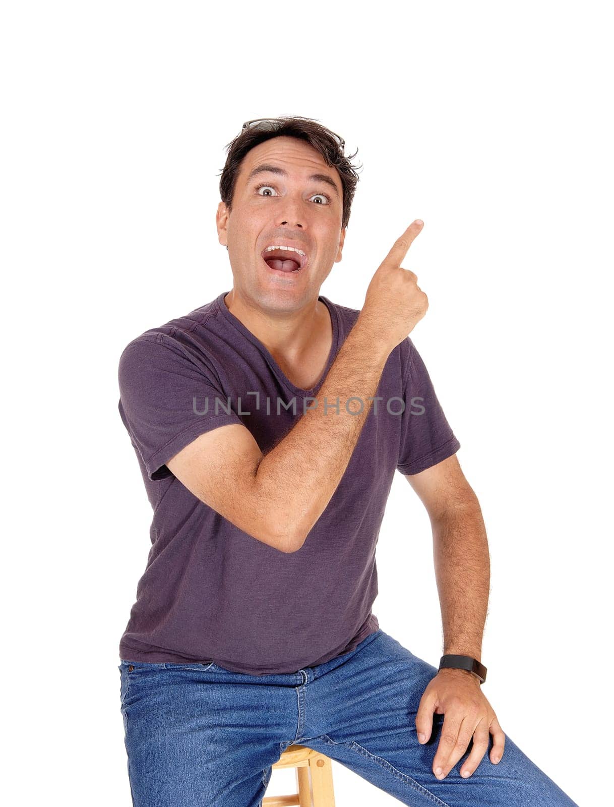 A young very handsome tall man sitting in jeans and a t-shirt with.his fingers pointing up and shouting, isolated for white background.