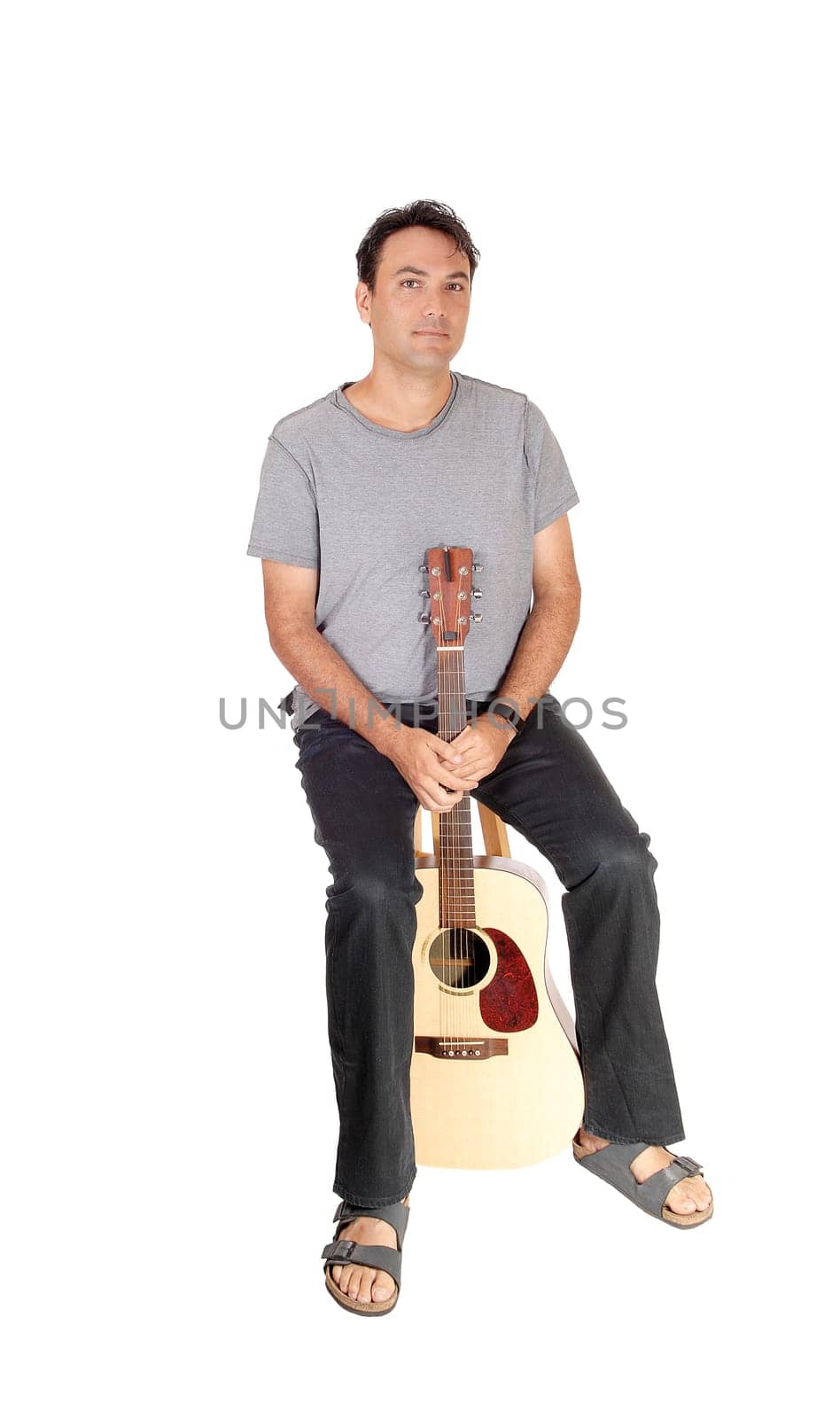 A young handsome man taking a brake from playing the guitar, sitting.in jeans gray shirt and sandals, isolated for white background.