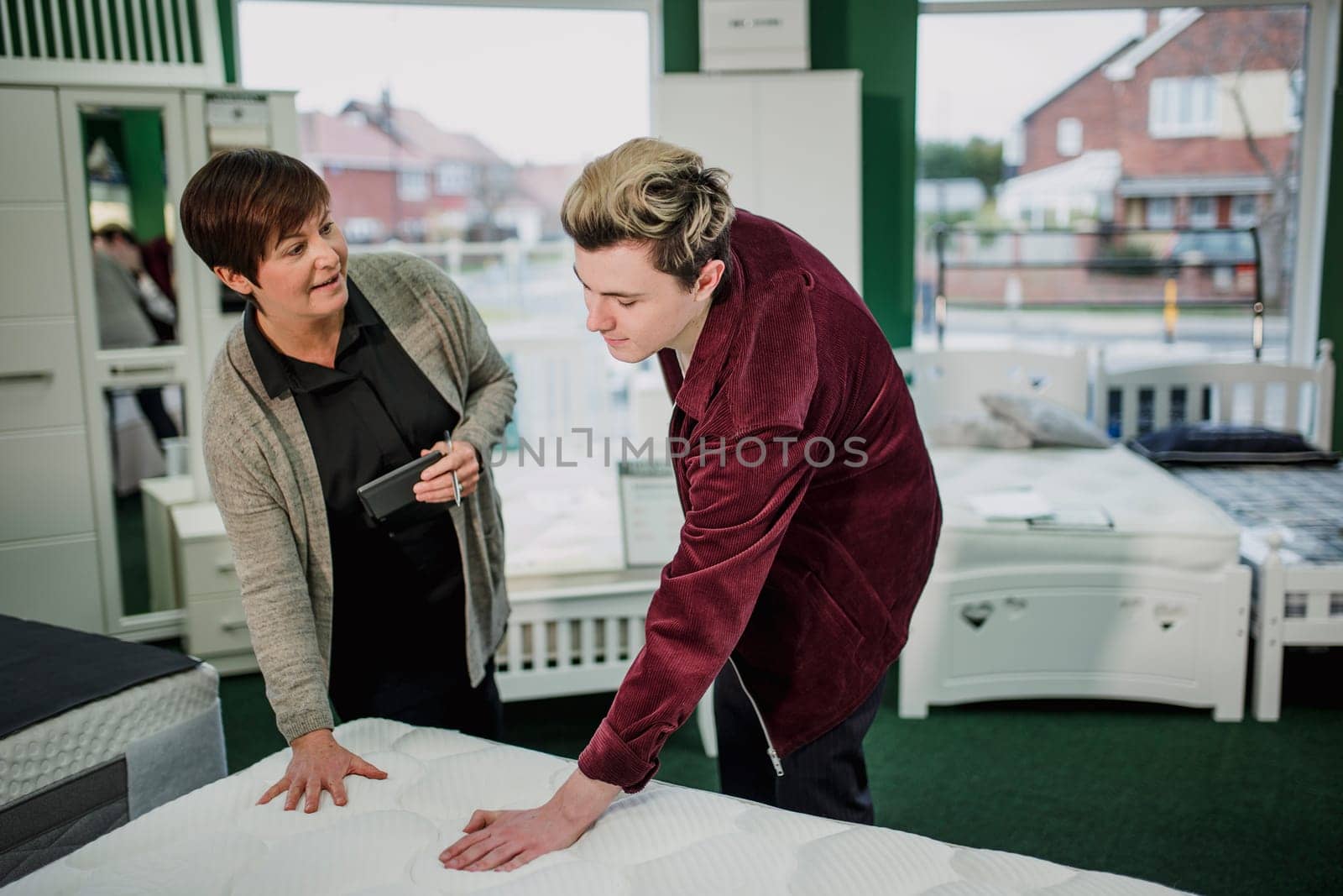 A saleswoman is assisting a young man as he shops for a new matress. She is talking to him while he tests a display.