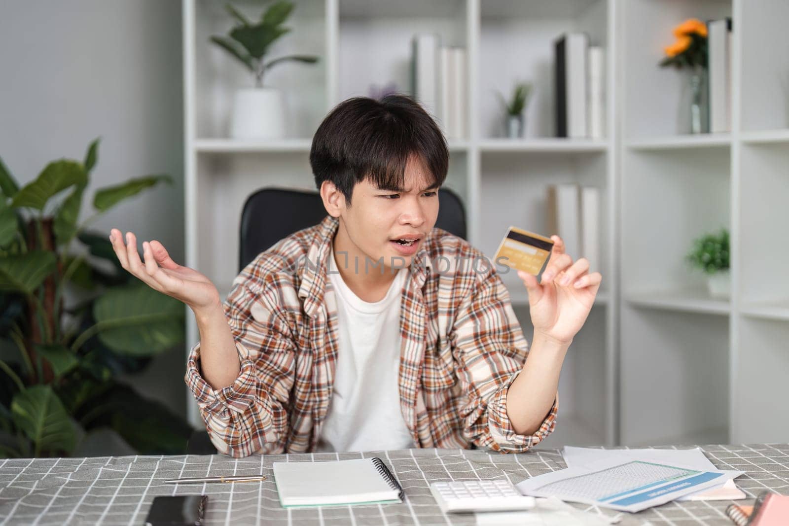 A young man looks frustrated while holding a credit card, reflecting confusion over his finances in a modern home office.