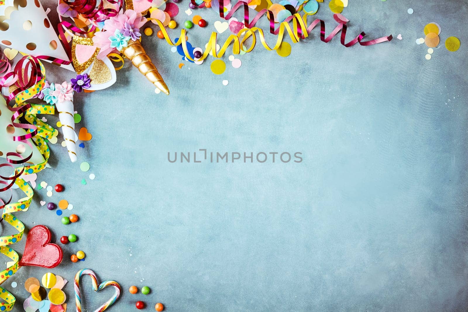 Colorful unicorn carnival border over a cool blue textured background with copy space with party hats, candy, streamers and hearts