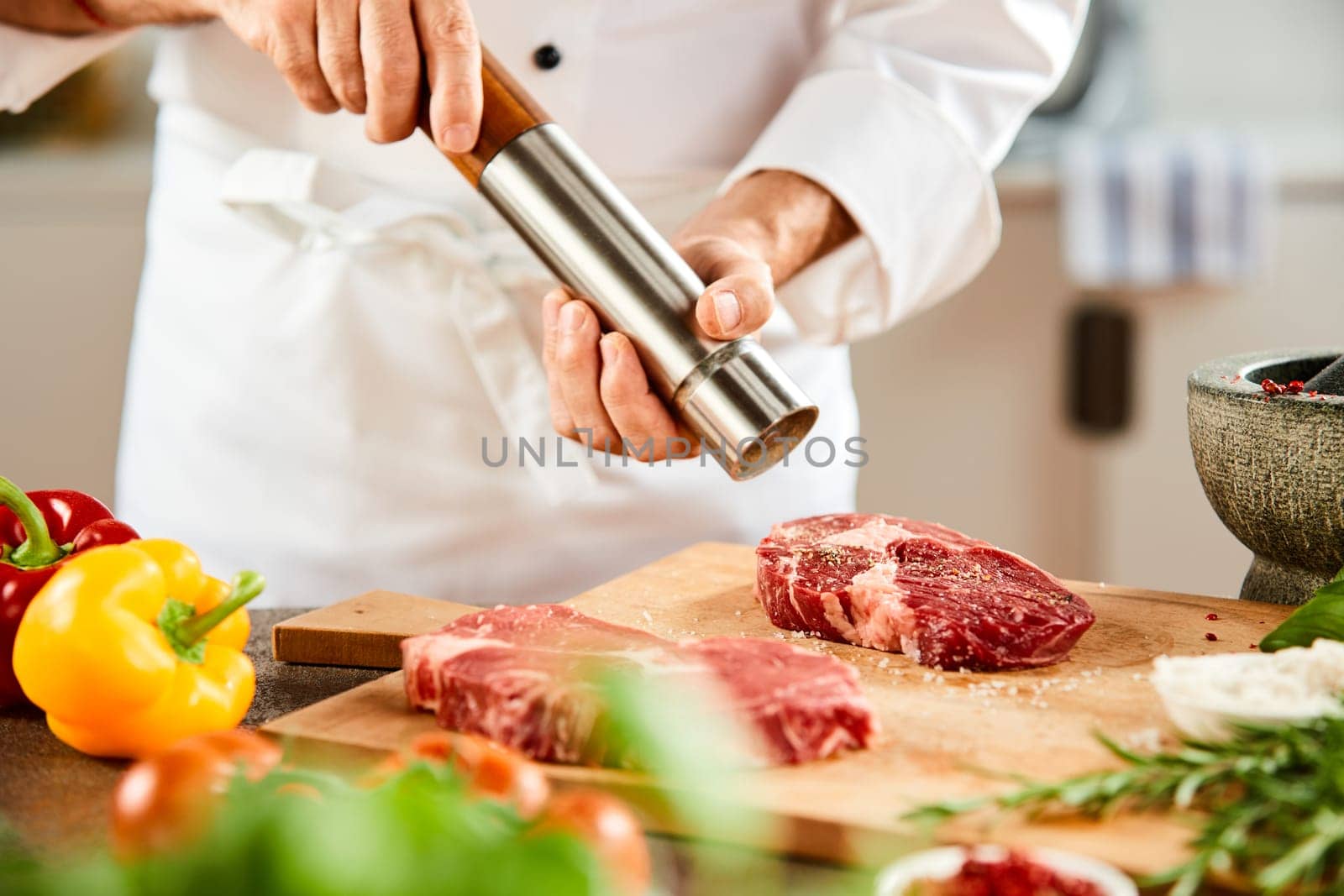 Side view of male chef in white jacket grinding pepper for salted meat net to vegetables in the foreground
