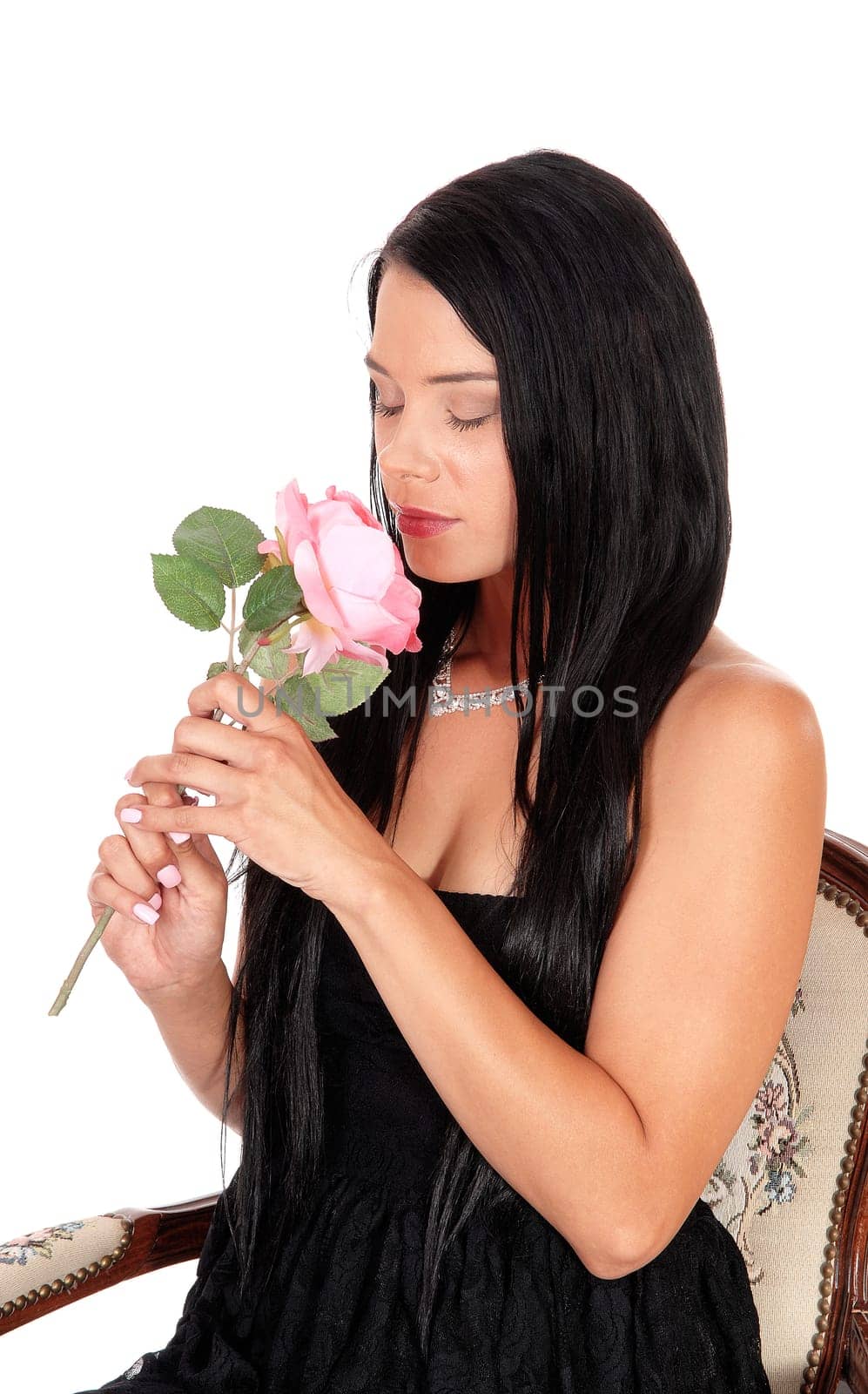 A beautiful young woman sitting in a black dress in an old armchair.holding a pink rose and smelling, isolated for white background.