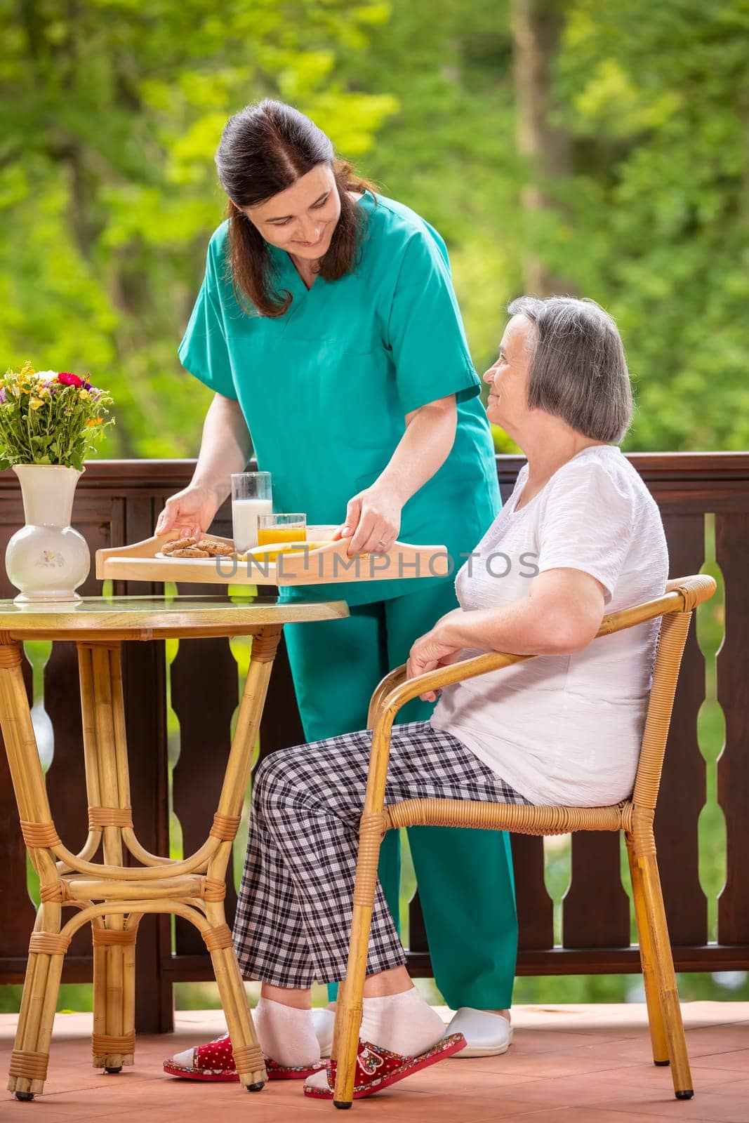 Happy nurse or caregiver serving healthy breakfast to elderly woman on the terrace at nursing home.