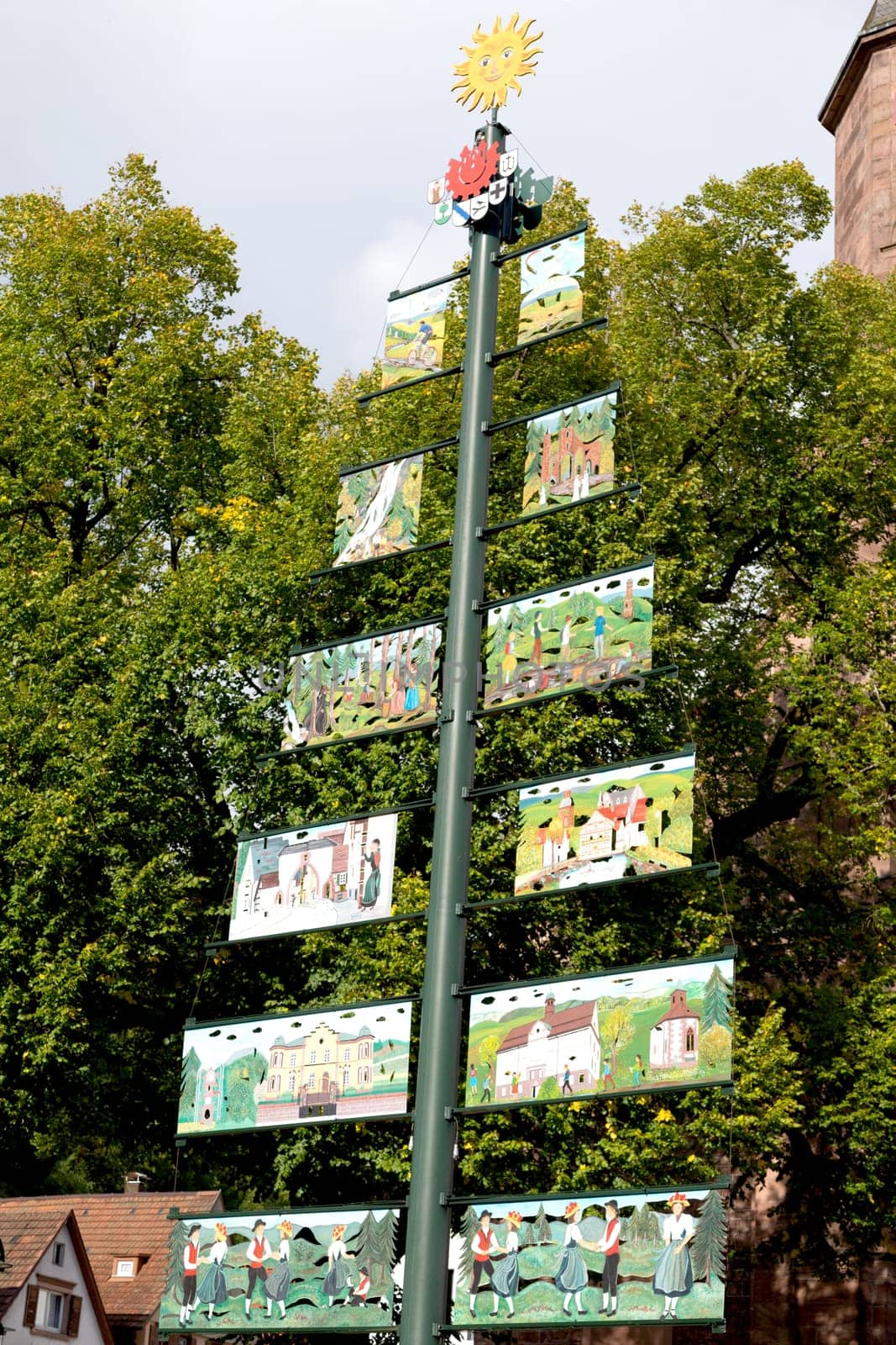 Large tree shaped signpost with pictures connected to sides and yellow smiling sun on top. Forest and houses in background.