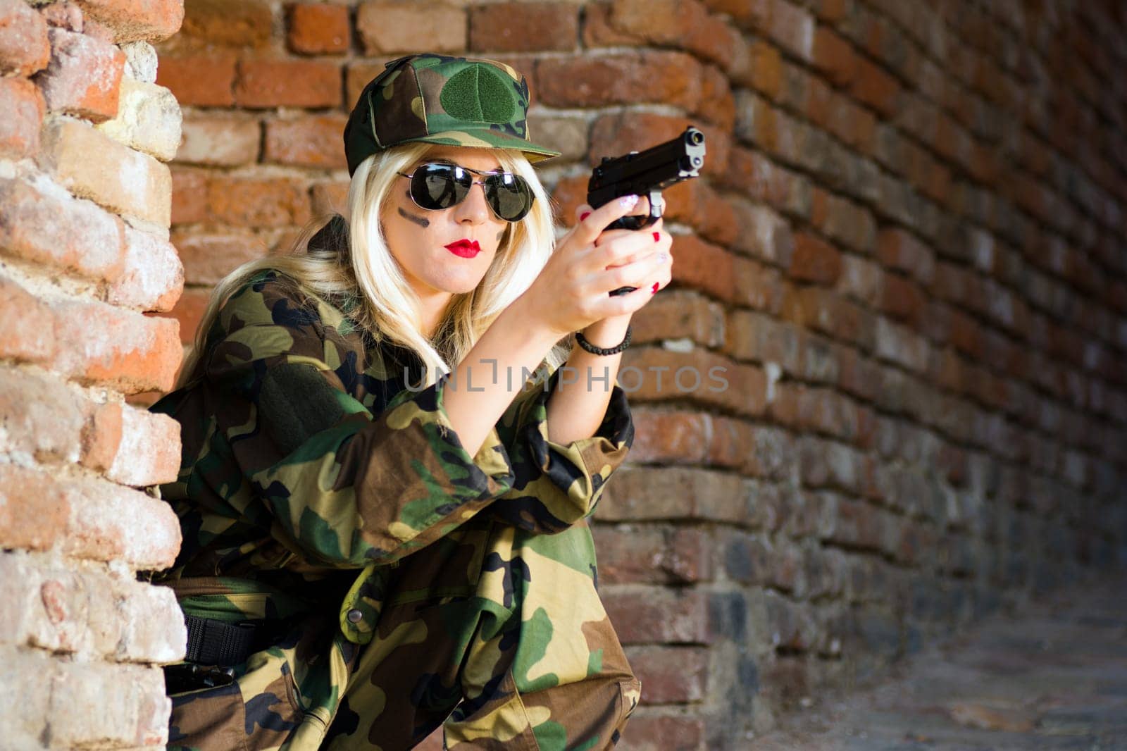 The female soldier with blond hair is holding gun on brick wall.