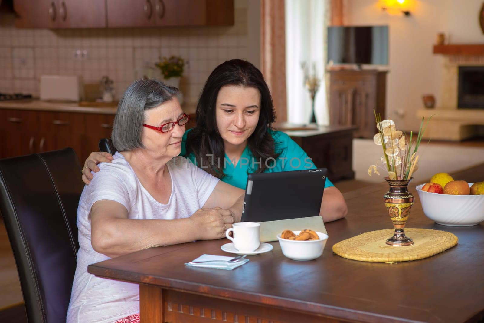Female nurse and senior woman using tablet PC in kitchen at nursing home.