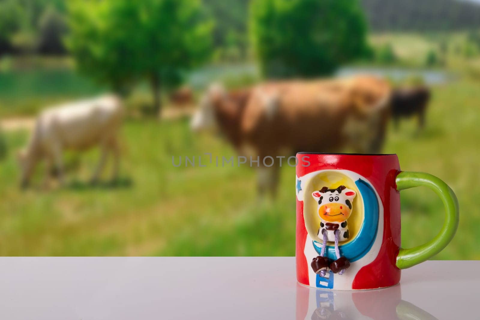 Funny cow as part of a milk cup on a bright table in front of blurred natural meadow with cows. Space for advertising your dairy products. Concept health.
