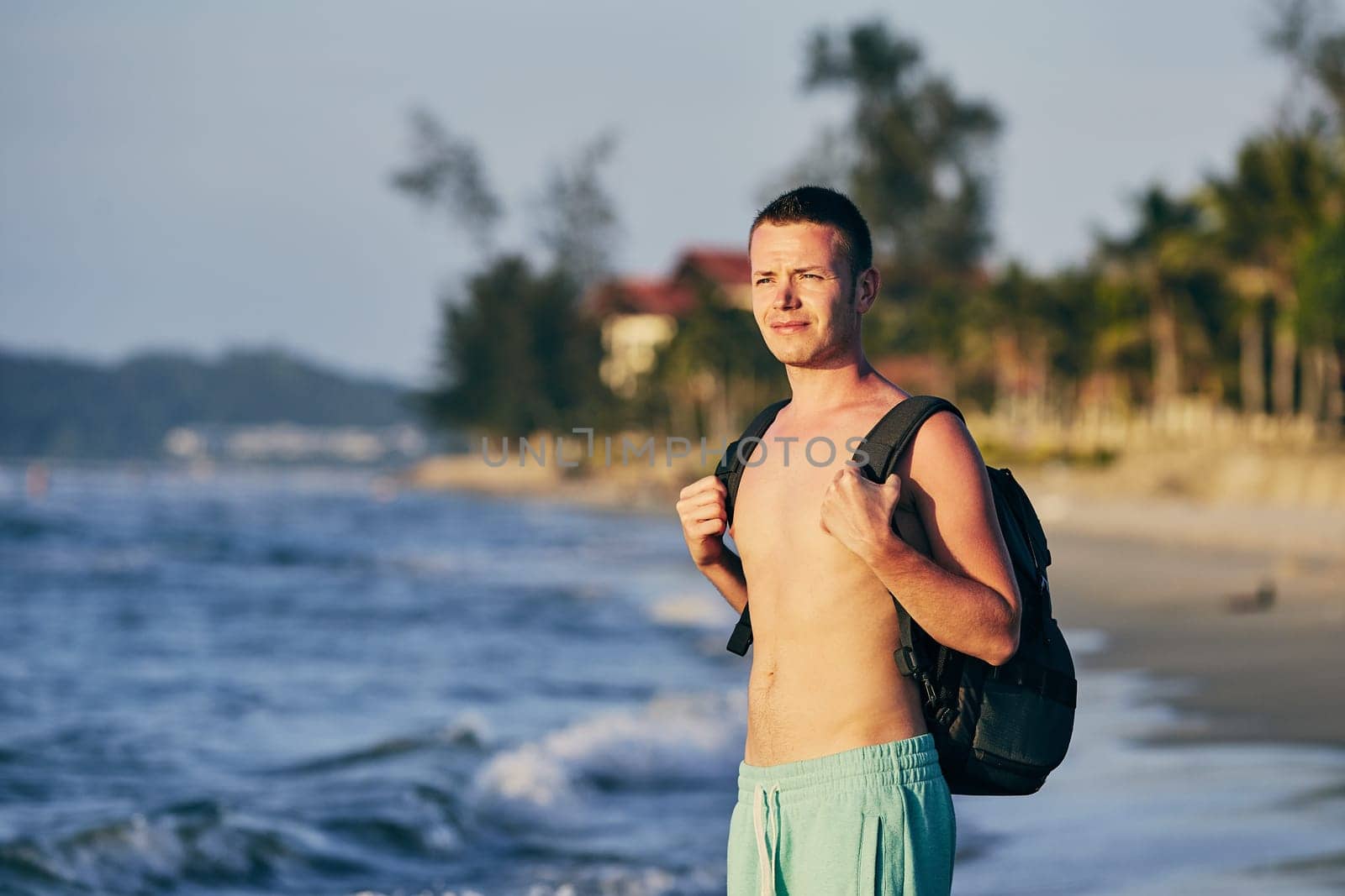 Young man (tourist) with backpack standing on sand beach.