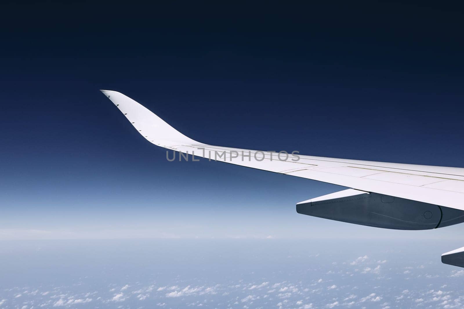 Wing of modern airplane flying over clouds against dark blue sky.