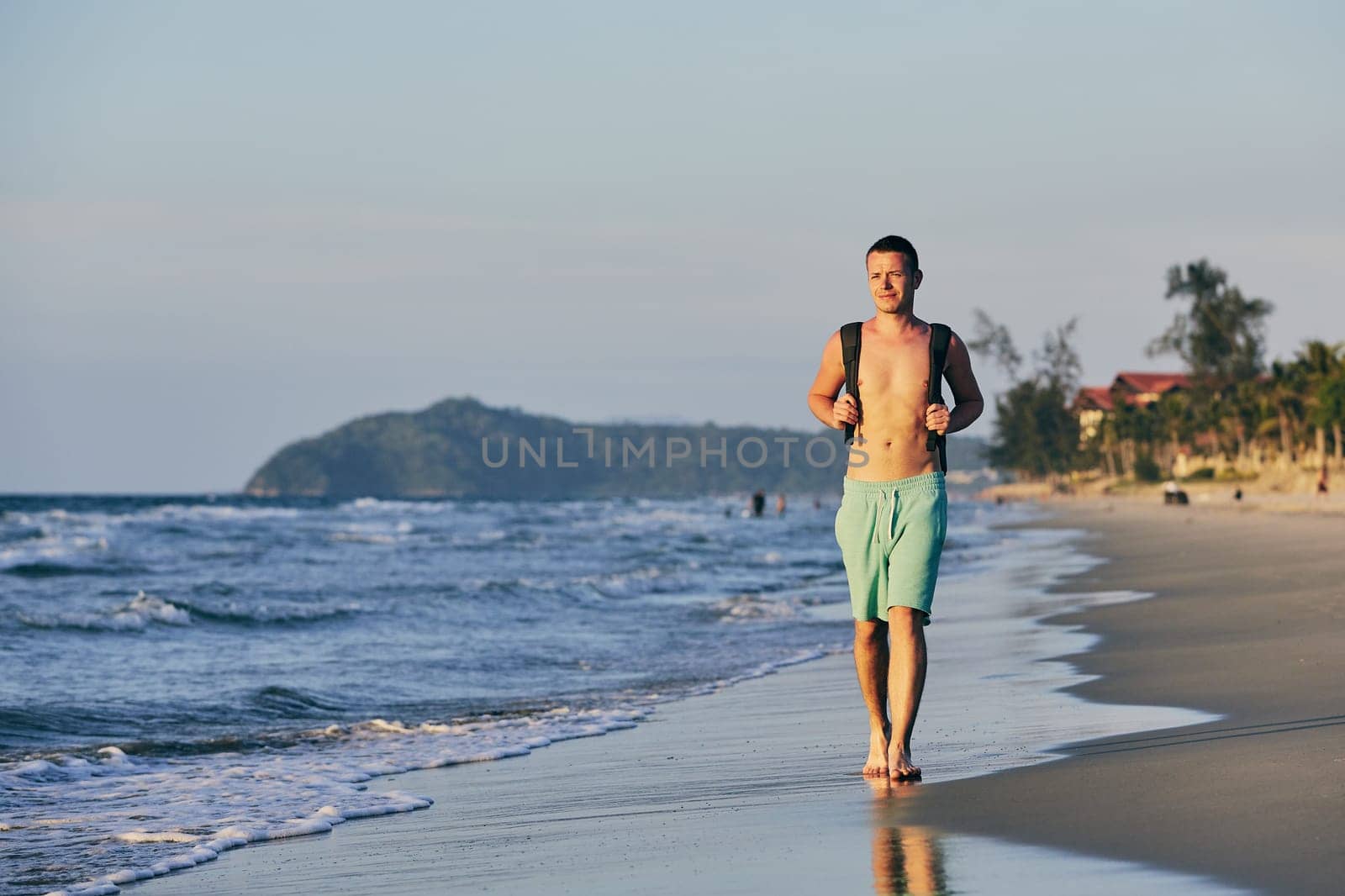 Young man (tourist) with backpack walking on sand beach.
