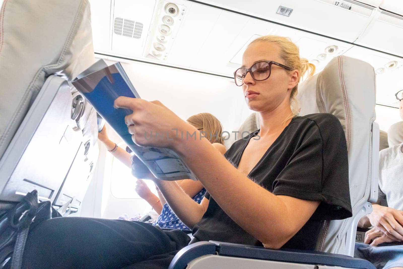 Cheerful woman reading magazine on airplane during flight. Female traveler reading seated in passanger cabin.