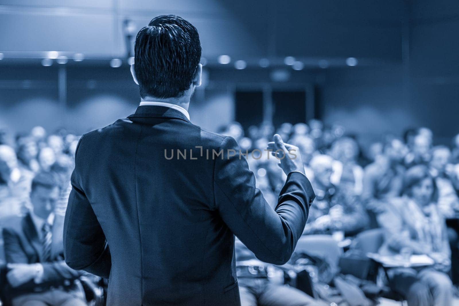 Speaker giving a talk on corporate business conference. Unrecognizable people in audience at conference hall. Business and Entrepreneurship event. Blue toned grayscale image.