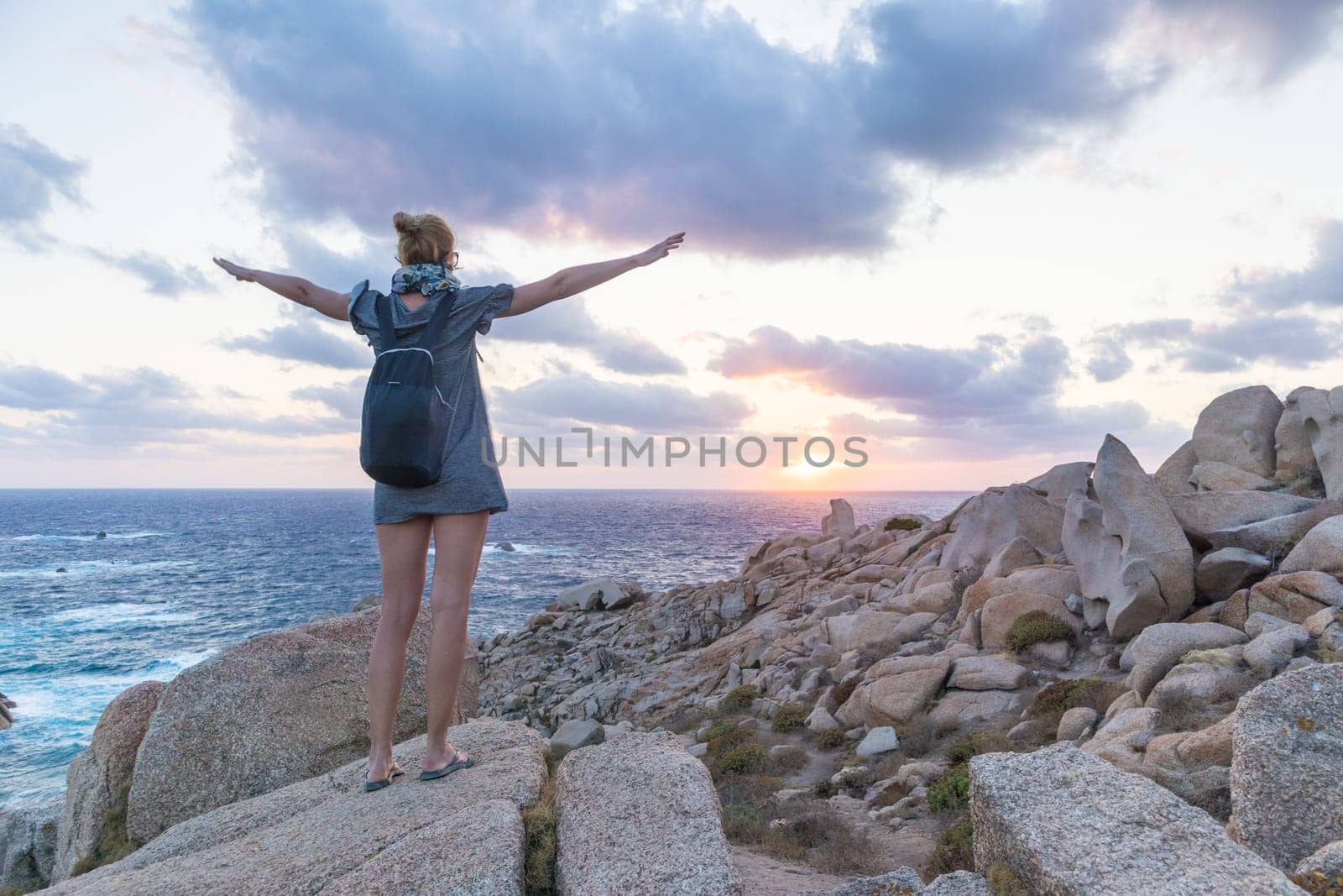 Female traveler, arms rised to the sky, watches a beautiful sunset on spectacular rocks of Capo Testa, Sardinia, Italy, a popular summer traveling destination in Europe.