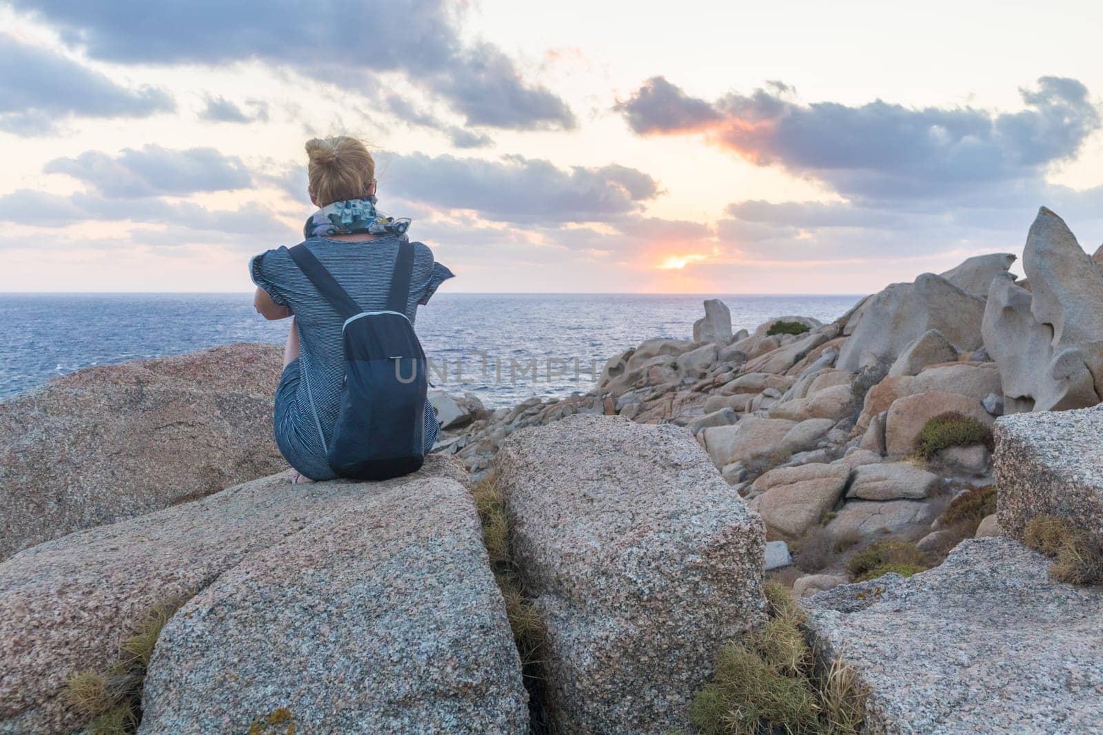Solitary young female traveler watches a beautiful sunset on spectacular rocks of Capo Testa, Sardinia, Italy, a popular summer traveling destination in Europe.