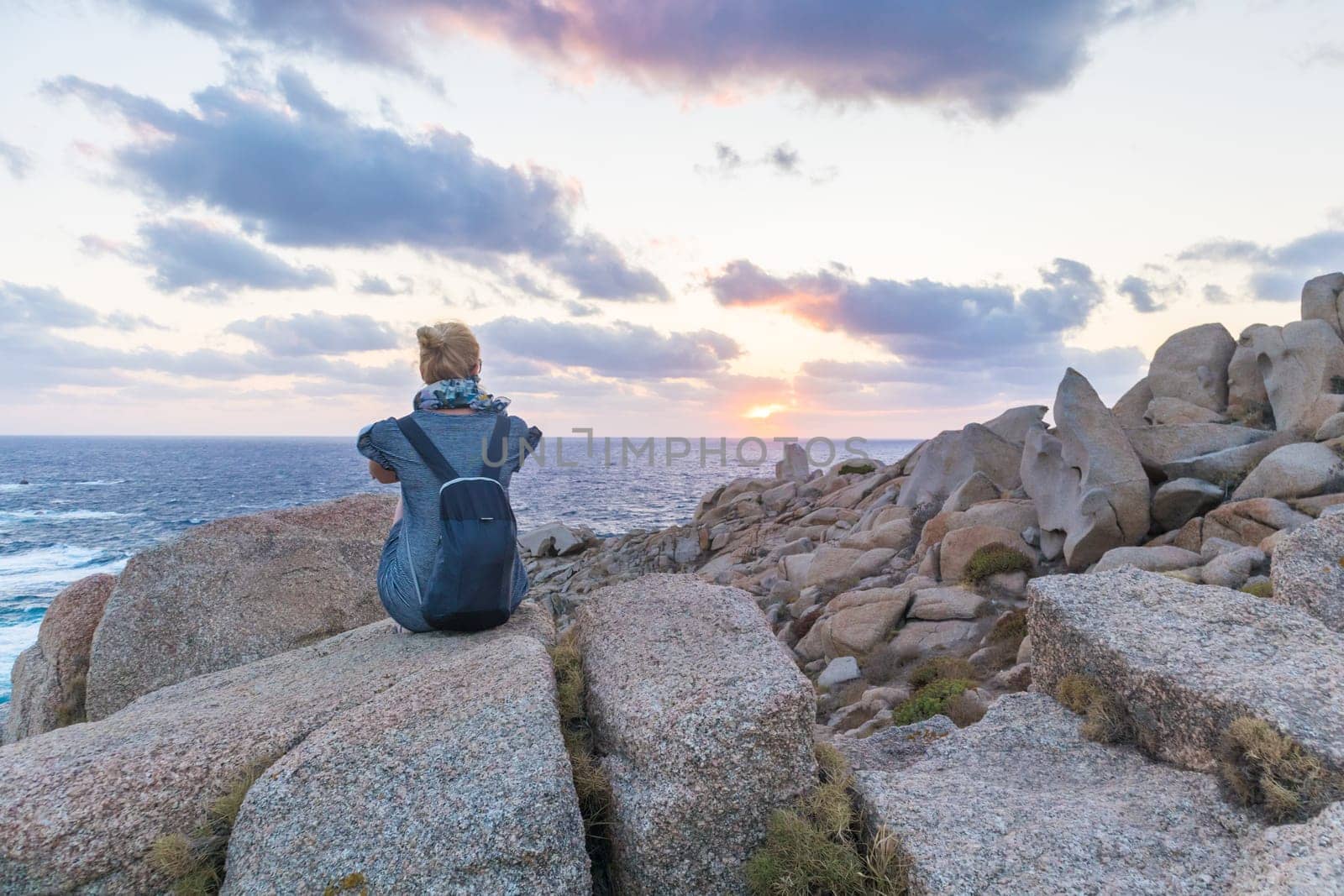 Solitary young female traveler watches a beautiful sunset on spectacular rocks of Capo Testa, Sardinia, Italy, a popular summer traveling destination in Europe.