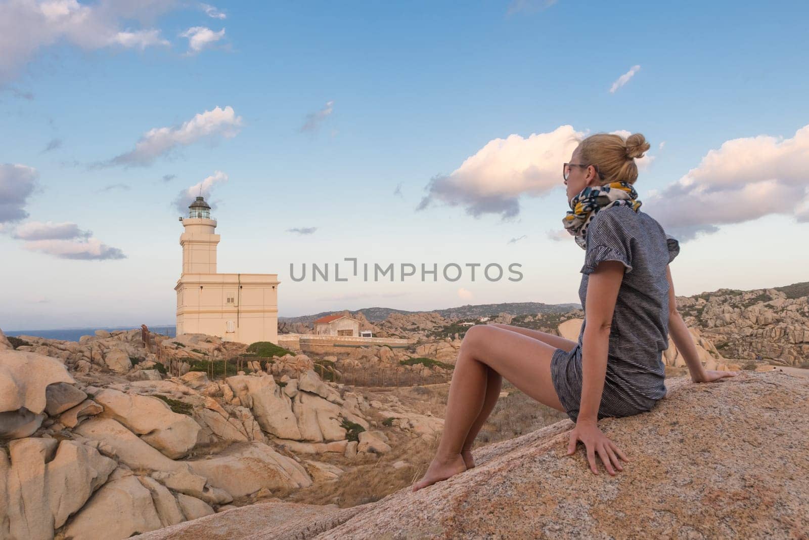 Solitary young female traveler watches a beautiful sunset over lighthouse and spectacular rock formations of Capo Testa, Sardinia, Italy, a popular summer traveling destination in Europe.