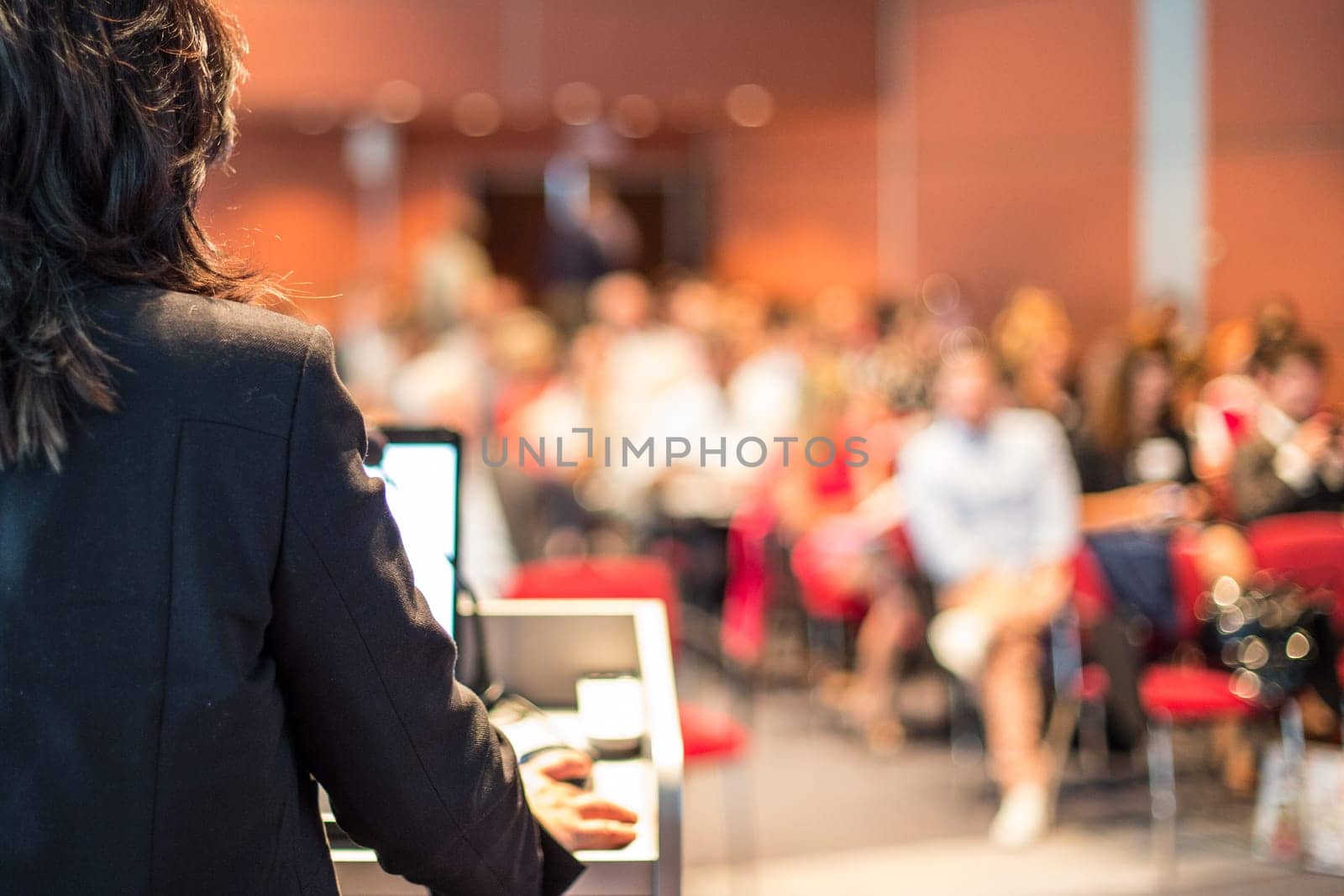Female speaker giving a talk on corporate business conference. Unrecognizable people in audience at conference hall. Business and Entrepreneurship event.
