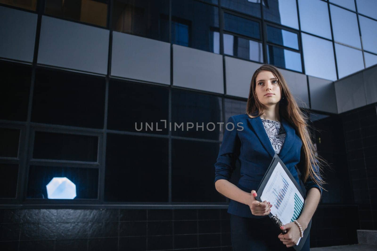 Successful beautiful young business woman standing against a background of buildings and holding folder.