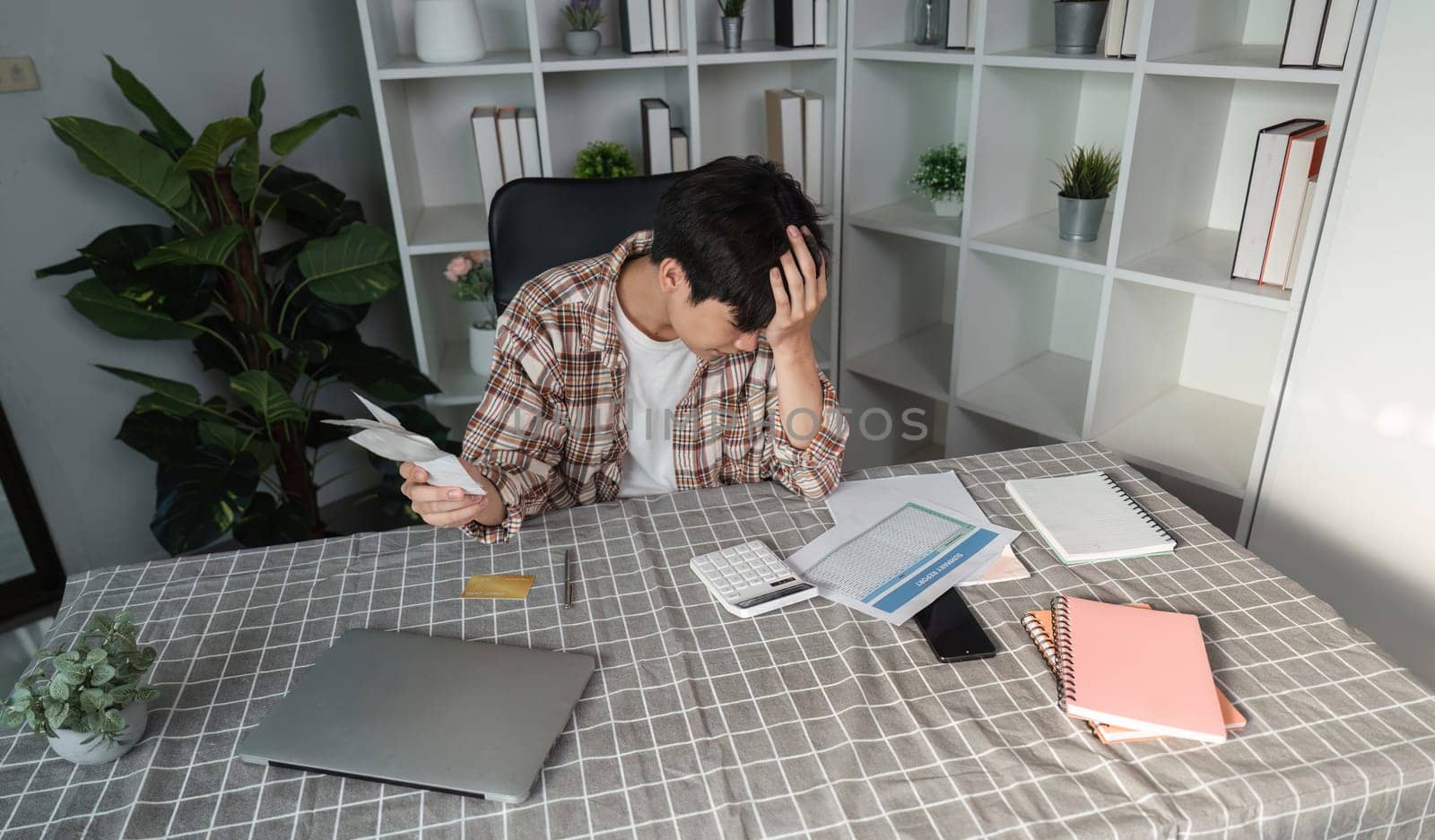 A young man appears stressed while managing his finances, using a calculator and phone amidst paperwork and notes.