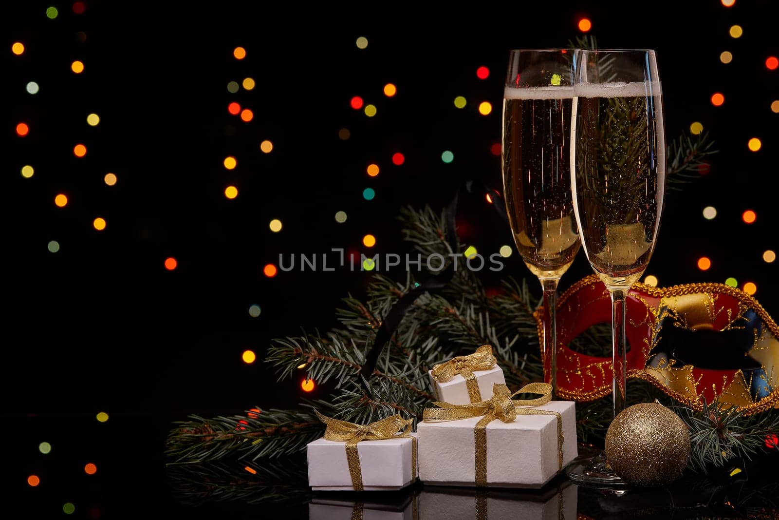 Two glasses with champange, fir tree branch with decoration, gift boxes on a black background with multi-colored lightes of garland. New year and Christmas.