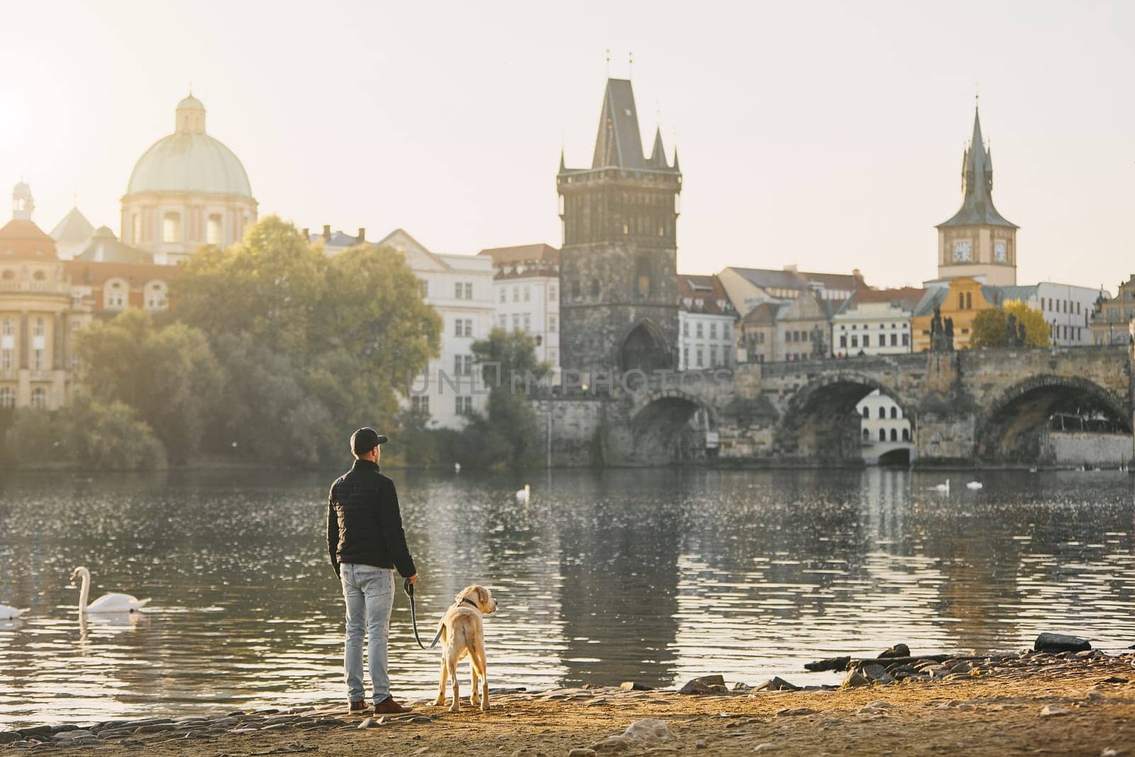 Morning walk with dog. Young man and his labrador retriever on riverbank against Old Town with Charles Bridge in Prague.