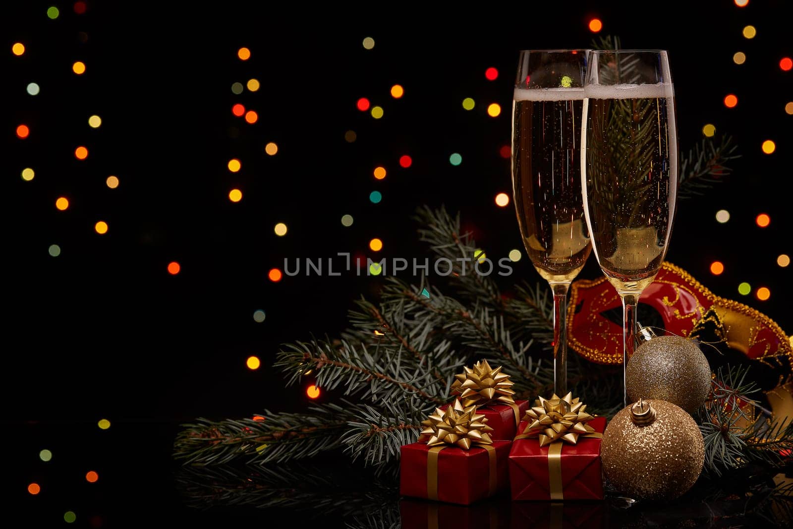 Two glasses with champange, fir tree branch with decoration, gift boxes on a black background with multi-colored lightes of garland. New year and Christmas.