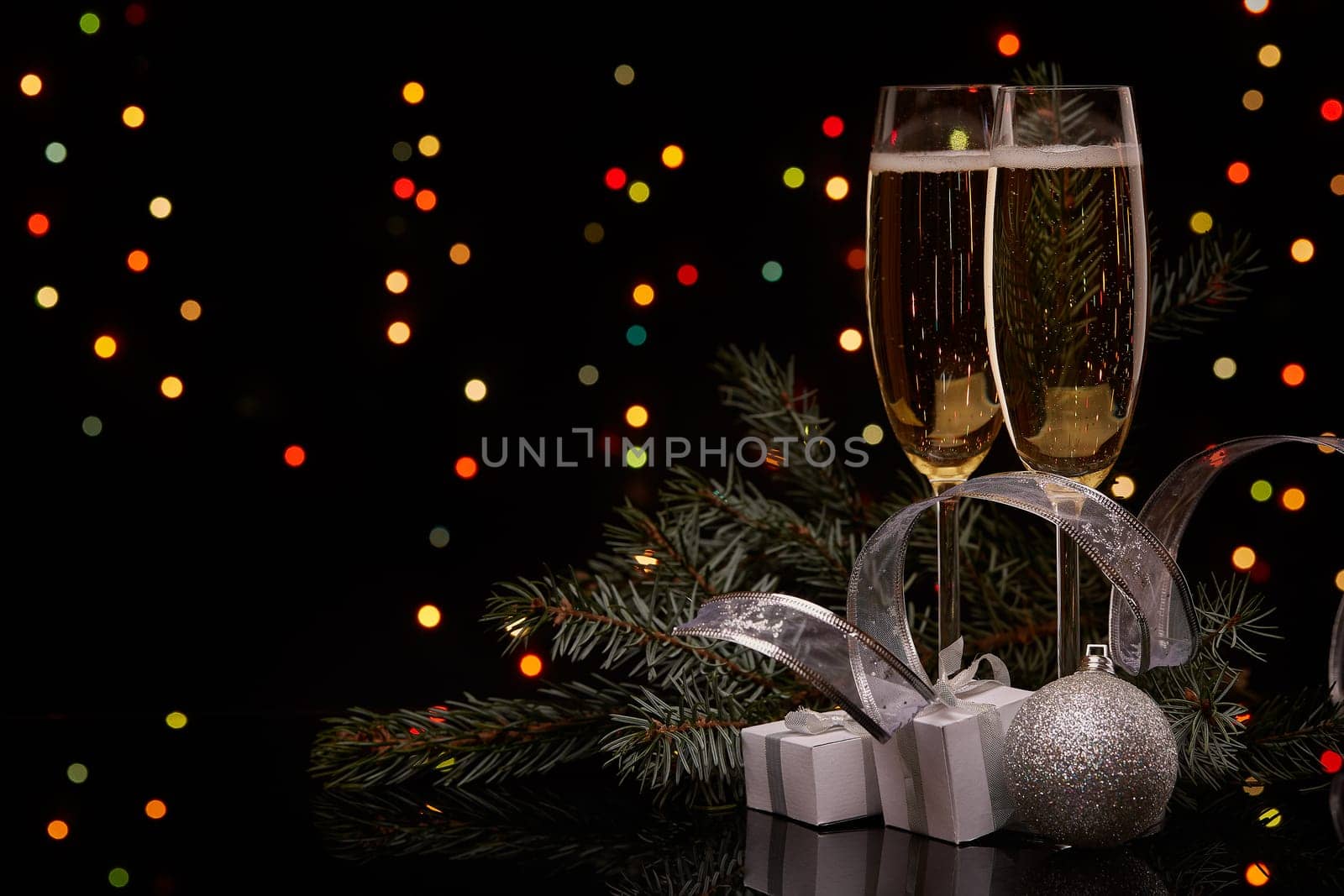 Two glasses with champange, fir tree branch with decoration, gift boxes on a black background with multi-colored lightes of garland. New year and Christmas.