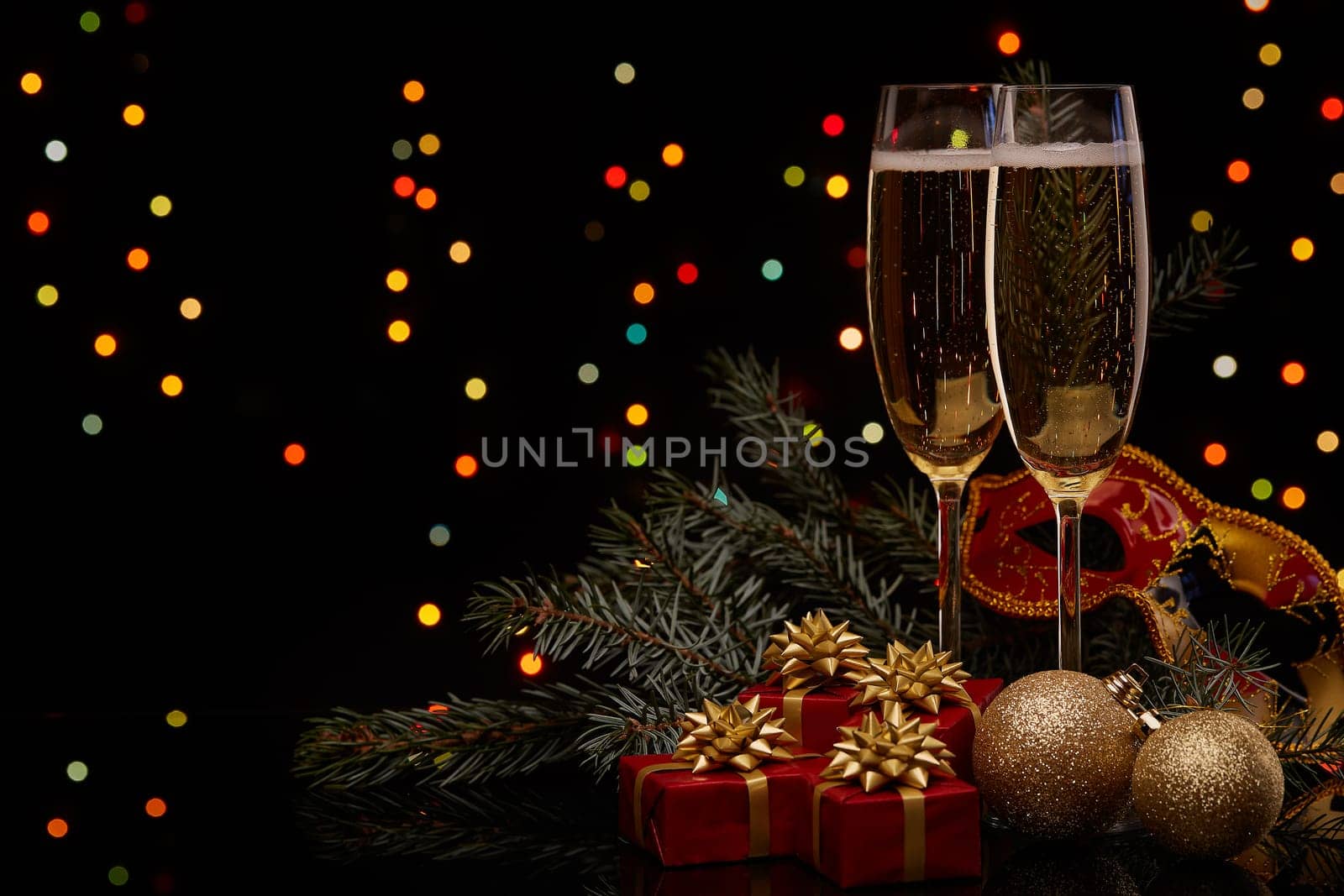 Two glasses with champange, fir tree branch with decoration, gift boxes on a black background with multi-colored lightes of garland. New year and Christmas.