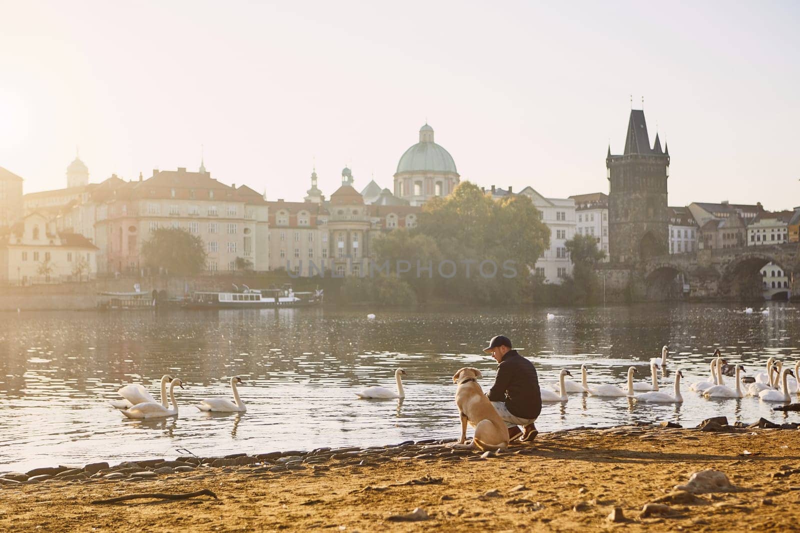 Morning walk with dog. Young man and his labrador retriever on riverbank against Old Town with Charles Bridge in Prague.