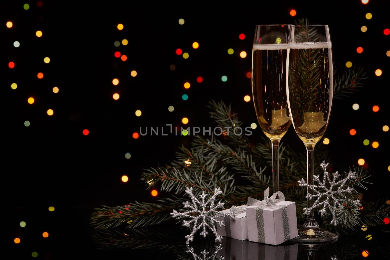 Two glasses with champange, fir tree branch with decoration, gift boxes on a black background with multi-colored lightes of garland. New year and Christmas.