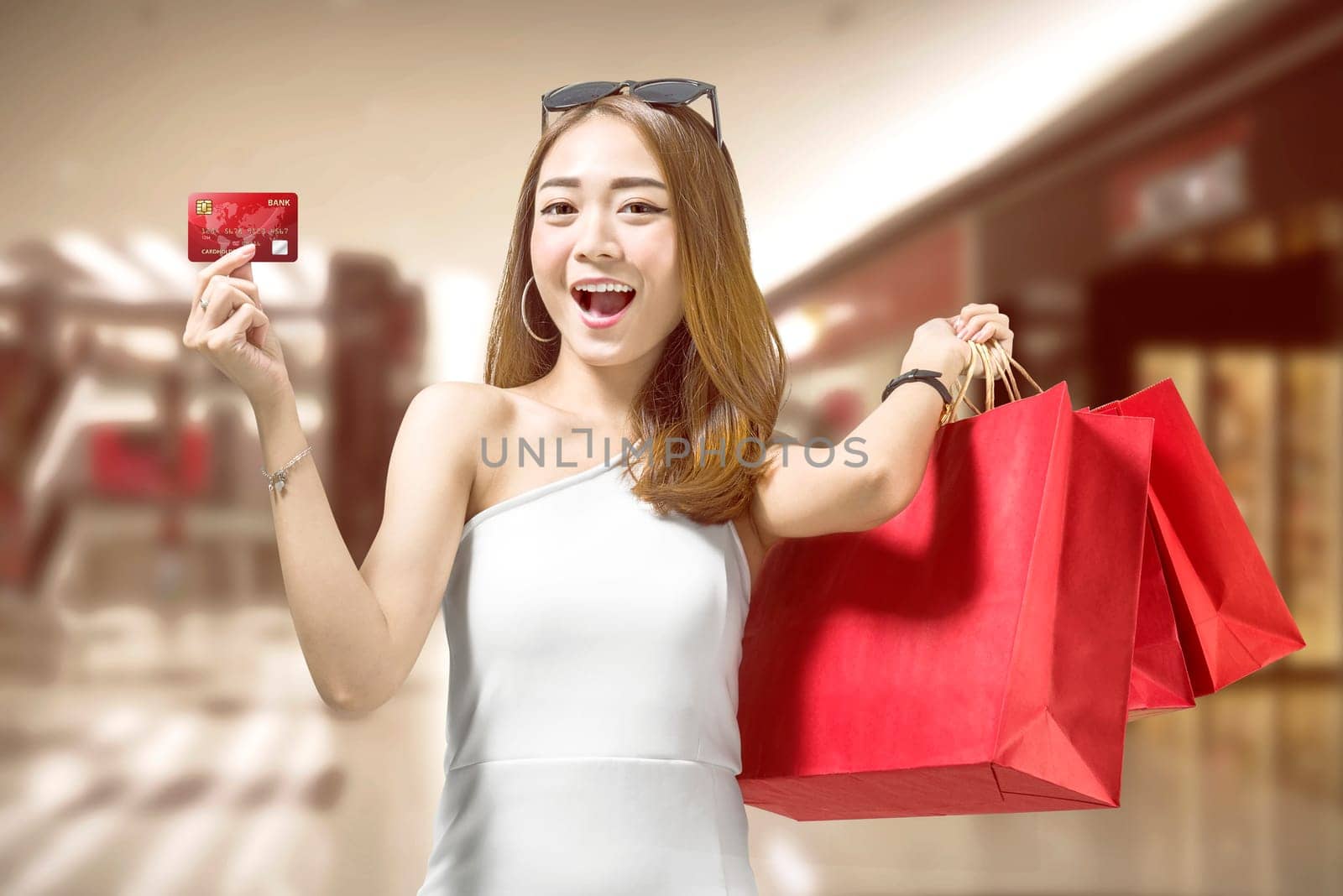 Cheerful asian woman with red shopping bags showing her credit card on the shopping mall