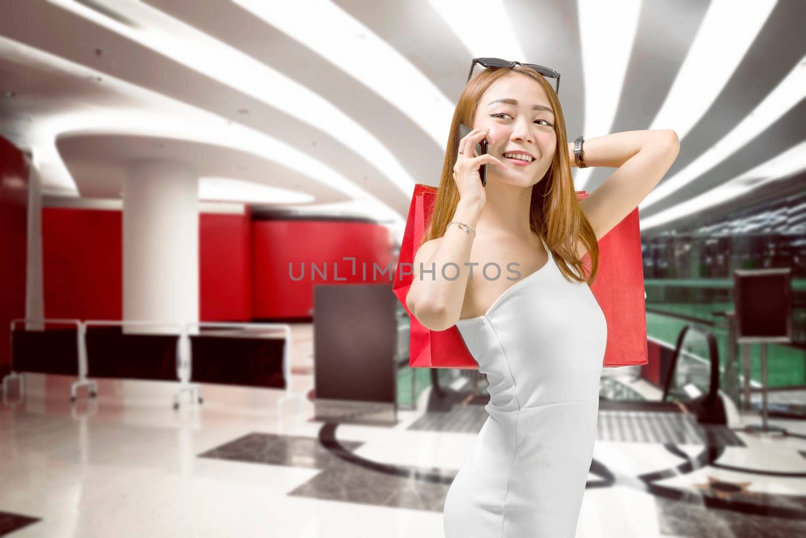 Smiling asian luxury woman with phone carrying shopping bag on the shopping mall