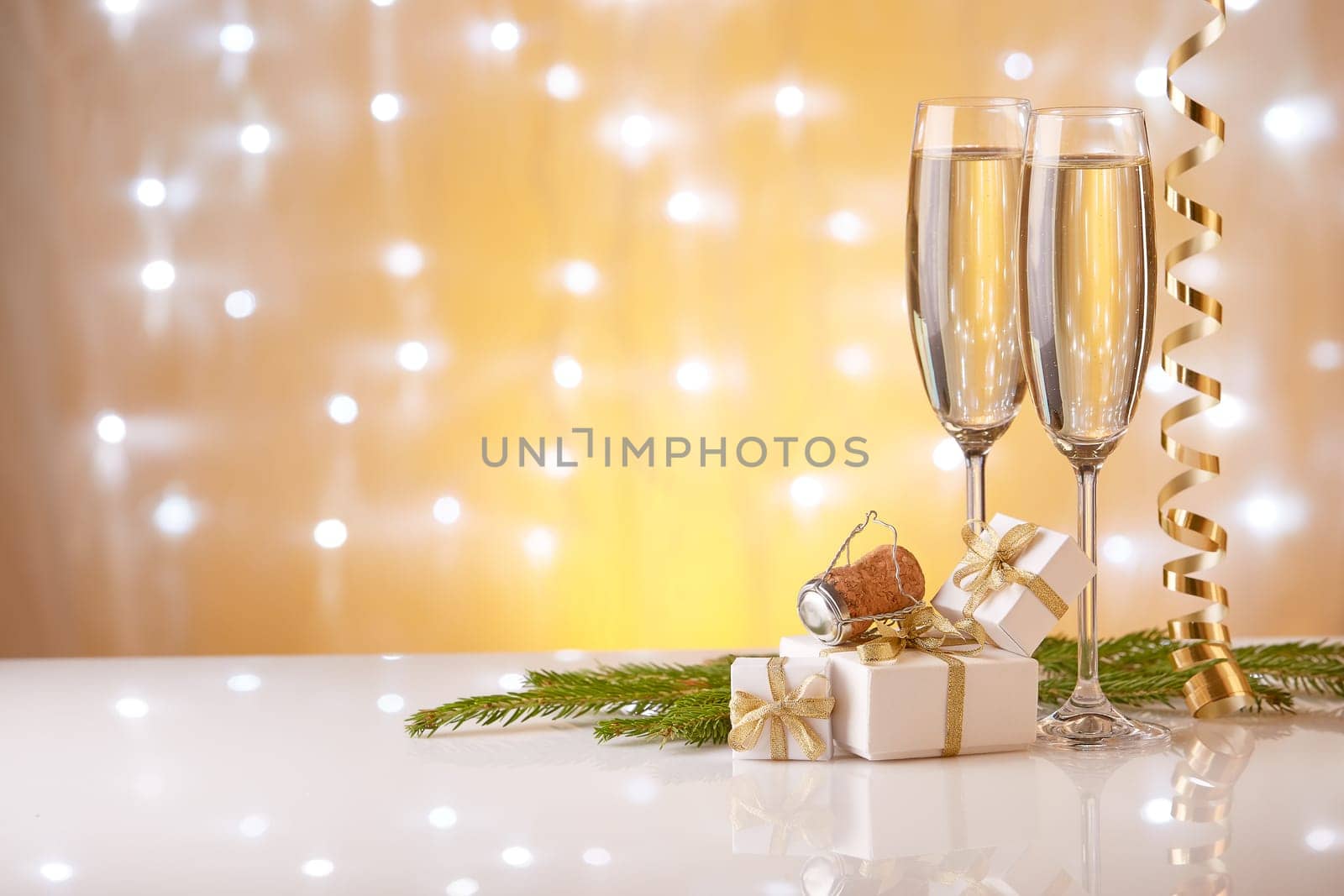 Two glasses with champange, fir tree branch, gift boxes and christmas decorations on a yellow background with lights of garland. New year and Christmas.
