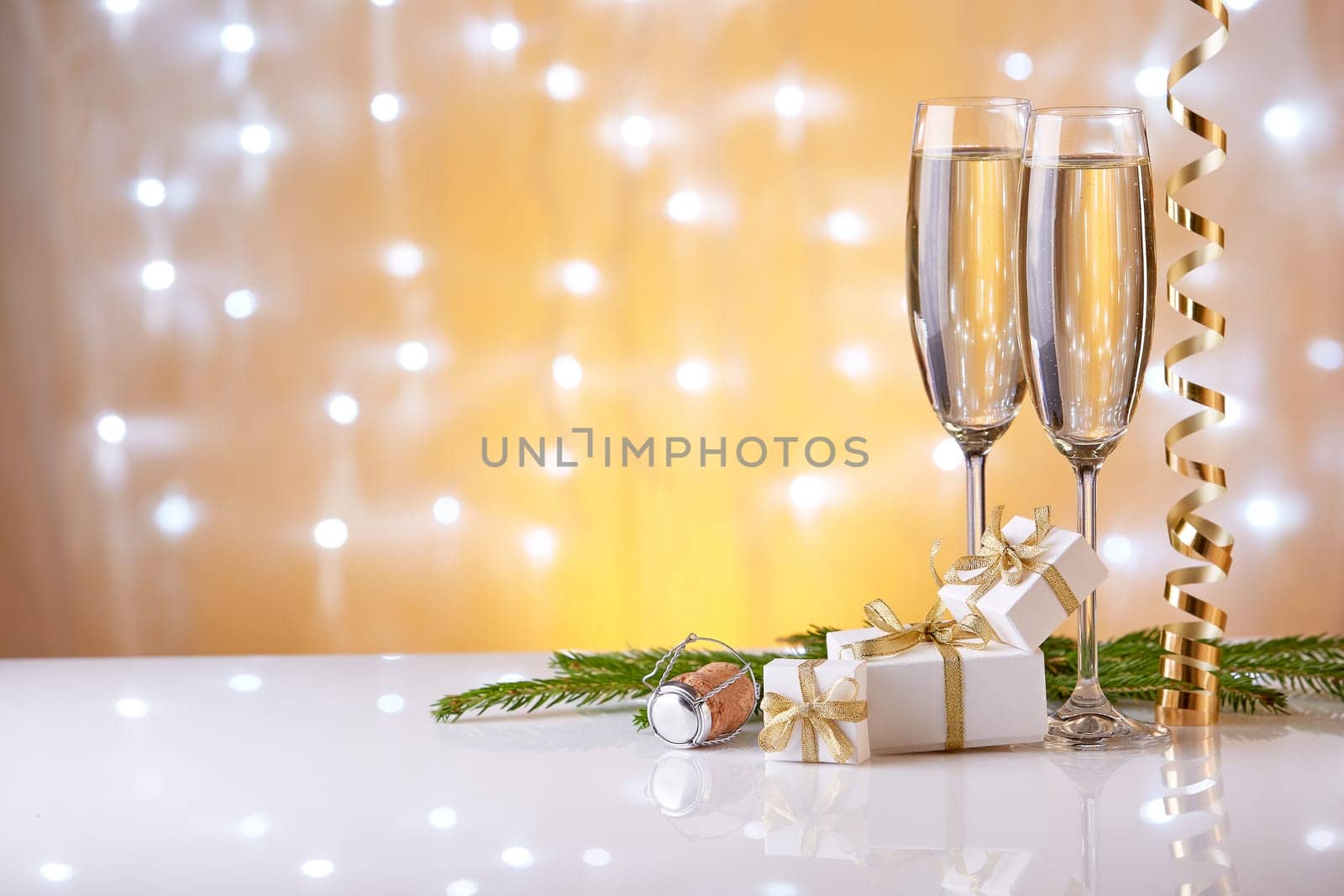 Two glasses with champange, fir tree branch, gift boxes and christmas decorations on a yellow background with lights of garland. New year and Christmas.