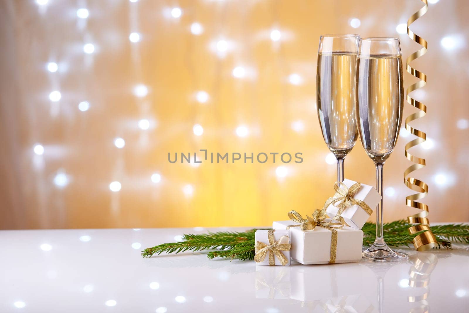 Two glasses with champange, fir tree branch, gift boxes and christmas decorations on a yellow background with lights of garland. New year and Christmas.