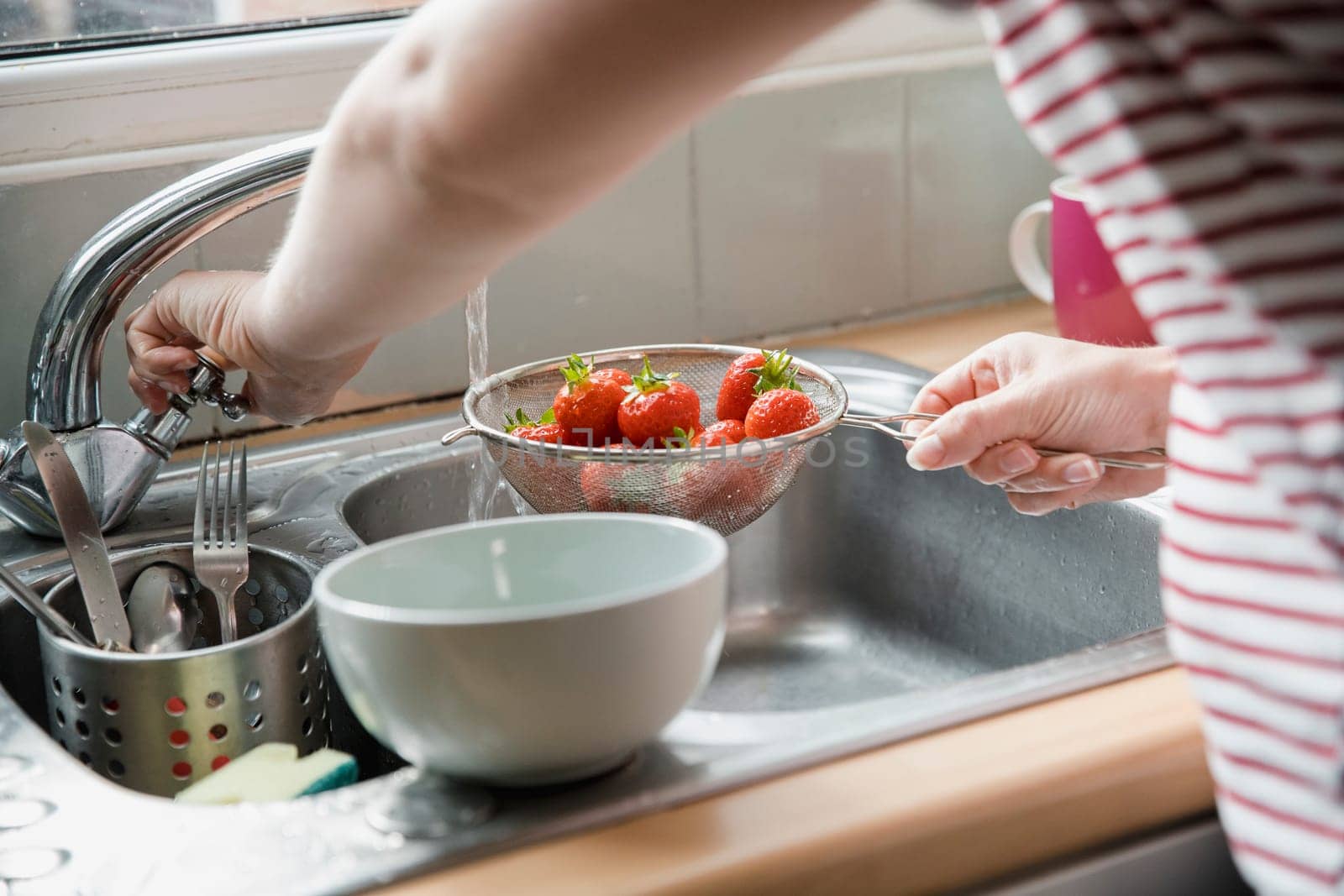 Close-up of an unrecognisable person standing at the kitchen sink rinsing a sieve full of strawberries with water.