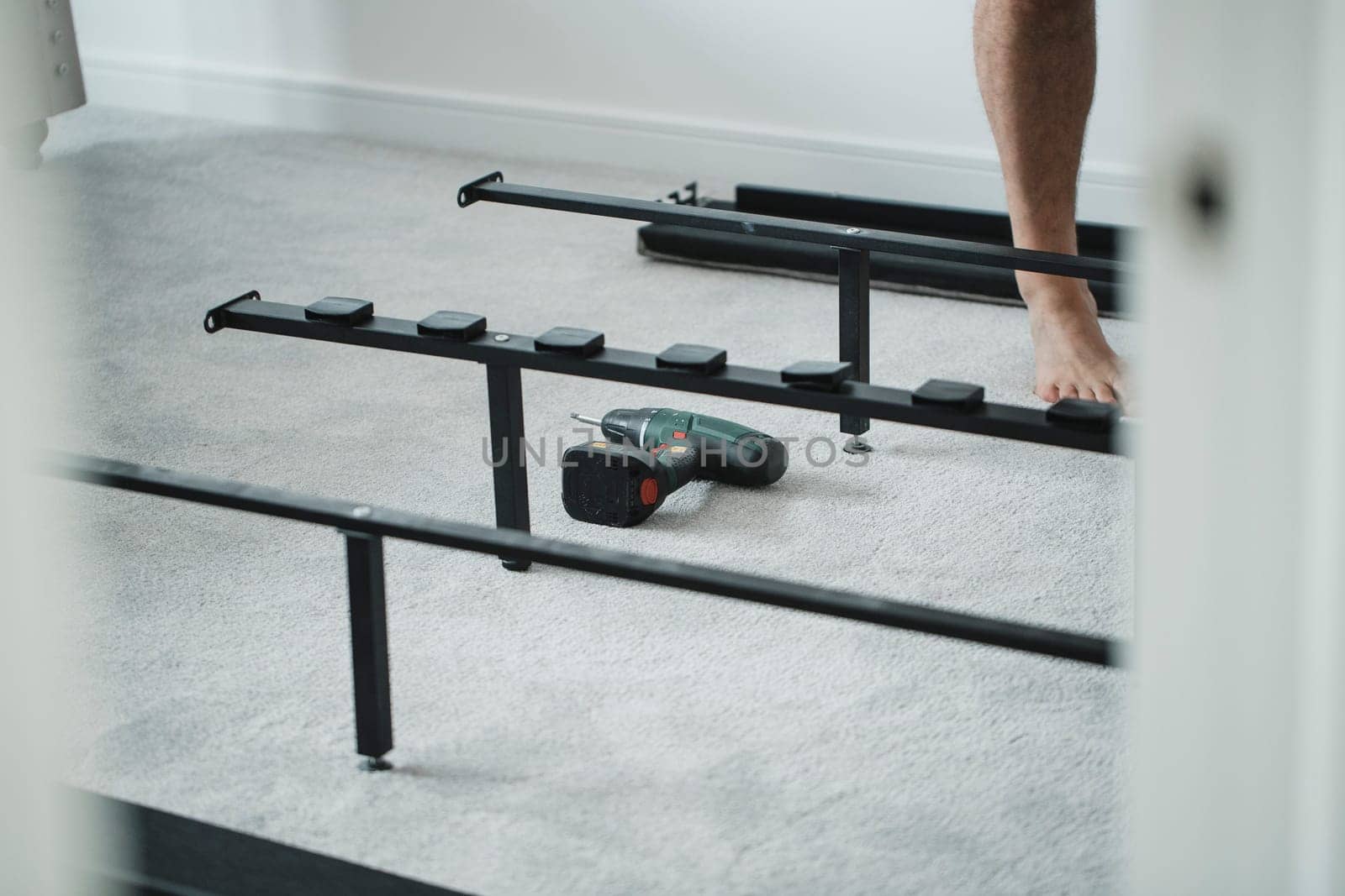A powerdrill is lying on the carpet near a bed frame which a man is putting together in a new home.