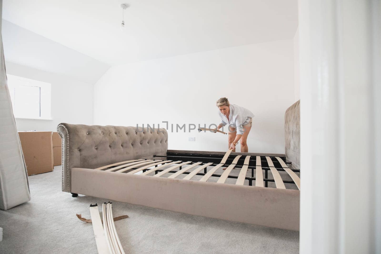 Mature woman is building her bed in her new home. She is placing wooden slats on the bed frame.
