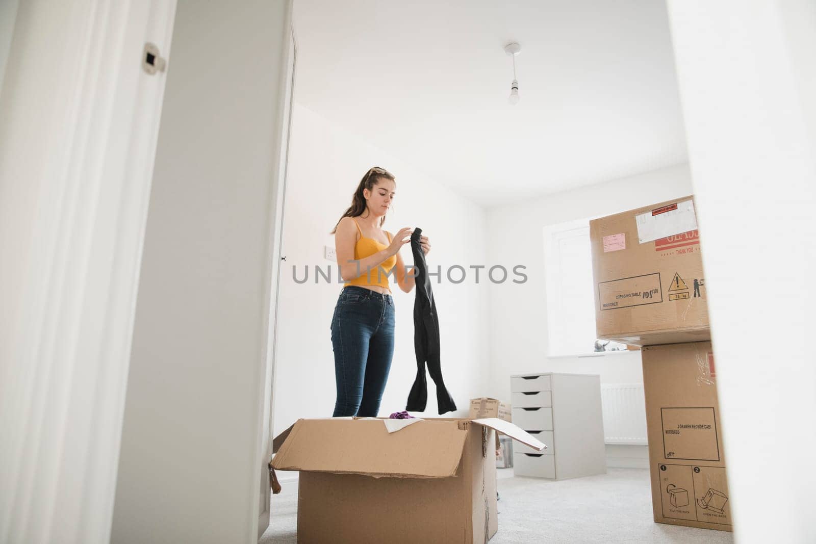 Teenage girl is unpacking in her new bedroom. She is folding clothes that she is taking out of a cardboard box.