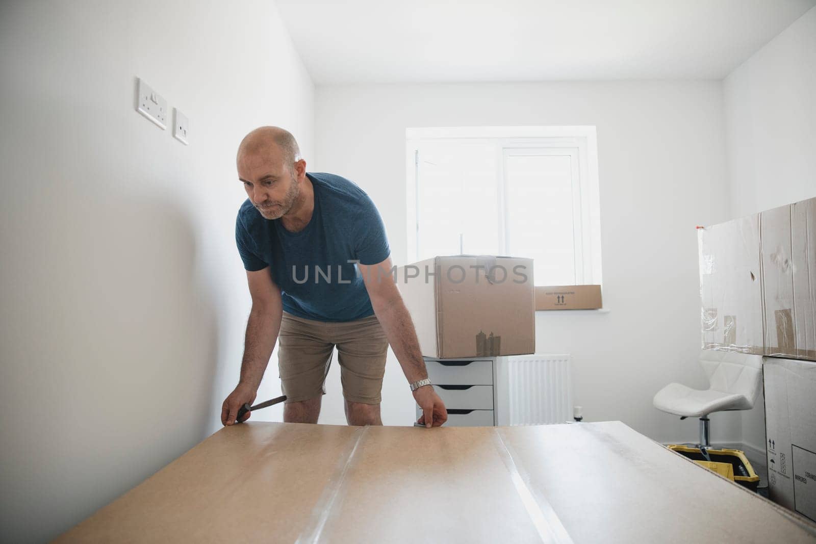 Mature man is moving furniture in to a new home. He is building a wardrobe in one of the bedrooms.