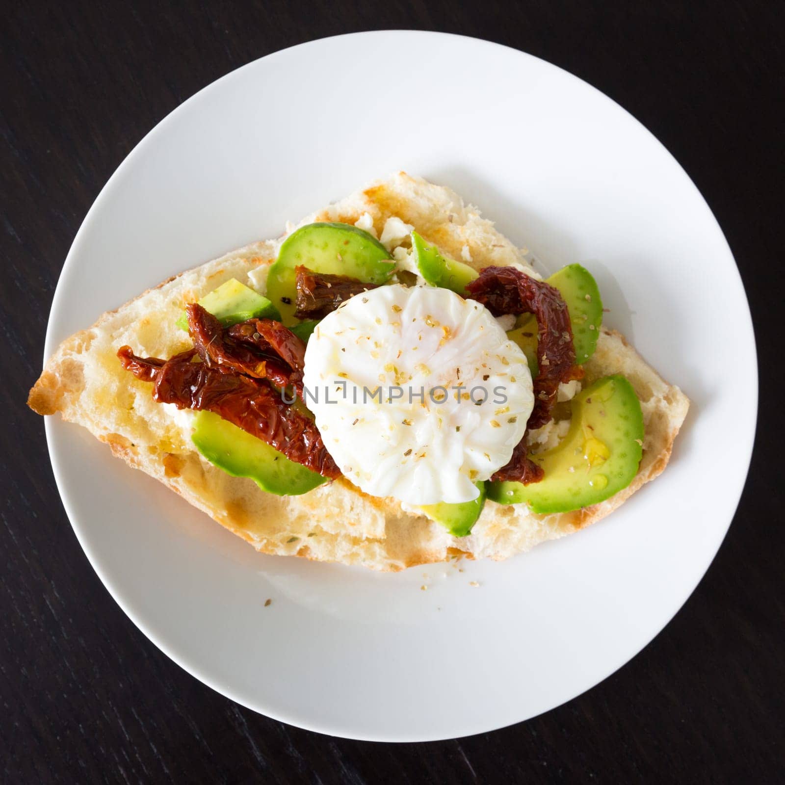 Poached egg on a toasted bun with avocados and dried tomatoes on white plate.