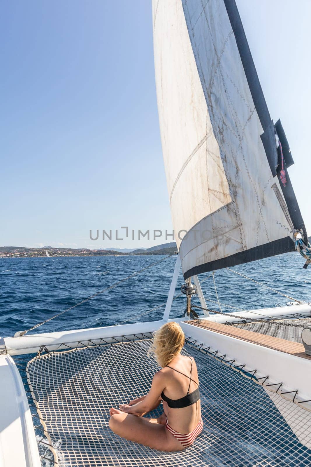 Woman in bikini tanning and relaxing on a summer sailin cruise, sitting on a luxury catamaran near picture perfect Palau town, Sardinia, Italy.