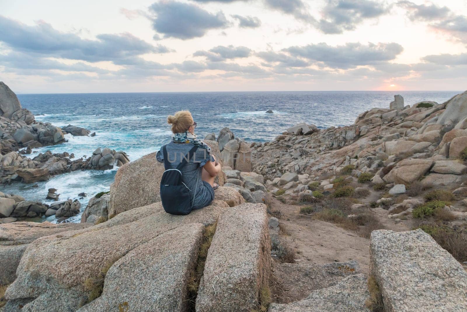 Solitary young female traveler watches a beautiful sunset on spectacular rocks of Capo Testa, Sardinia, Italy, a popular summer traveling destination in Europe.