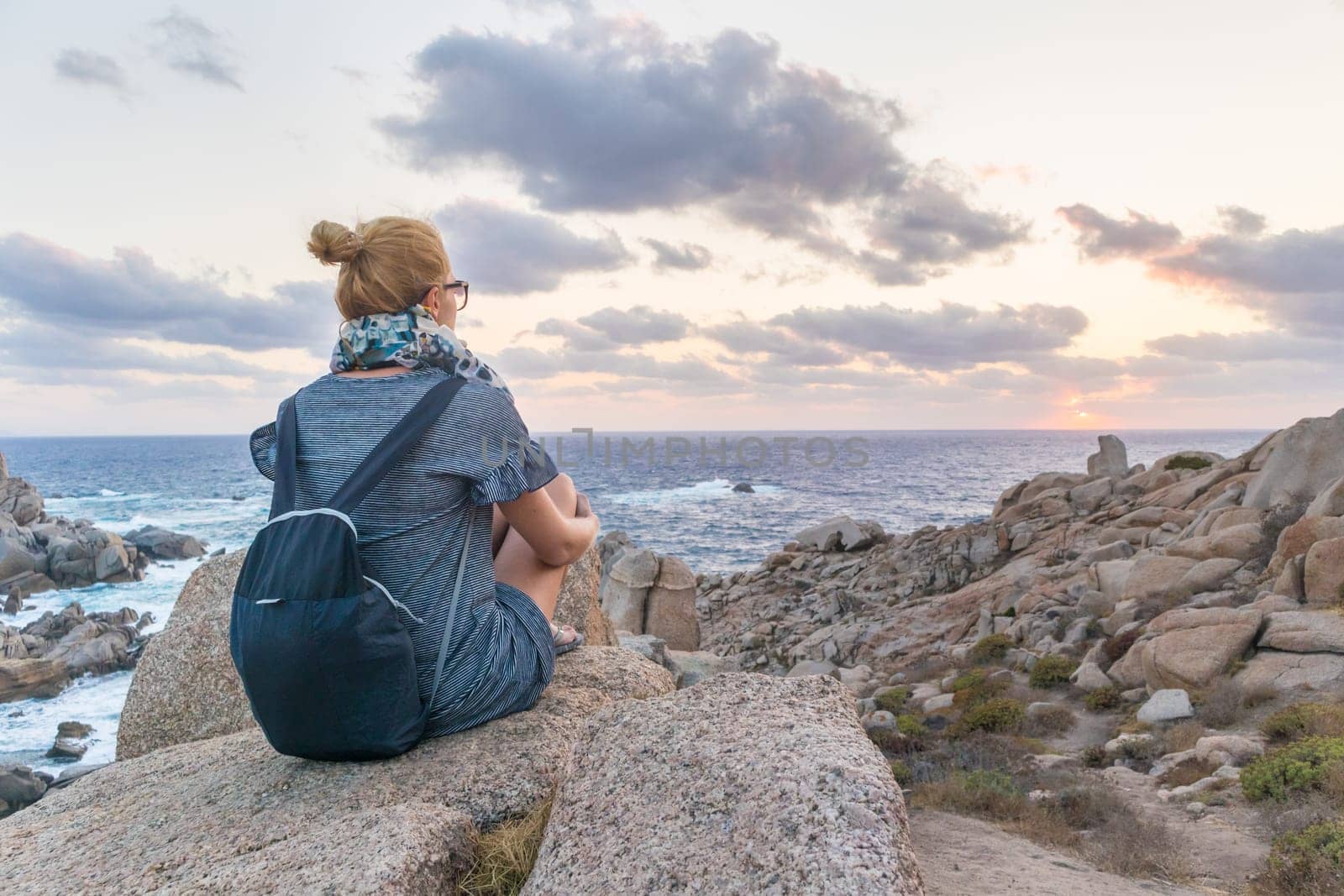 Solitary young female traveler watches a beautiful sunset on spectacular rocks of Capo Testa, Sardinia, Italy, a popular summer traveling destination in Europe.